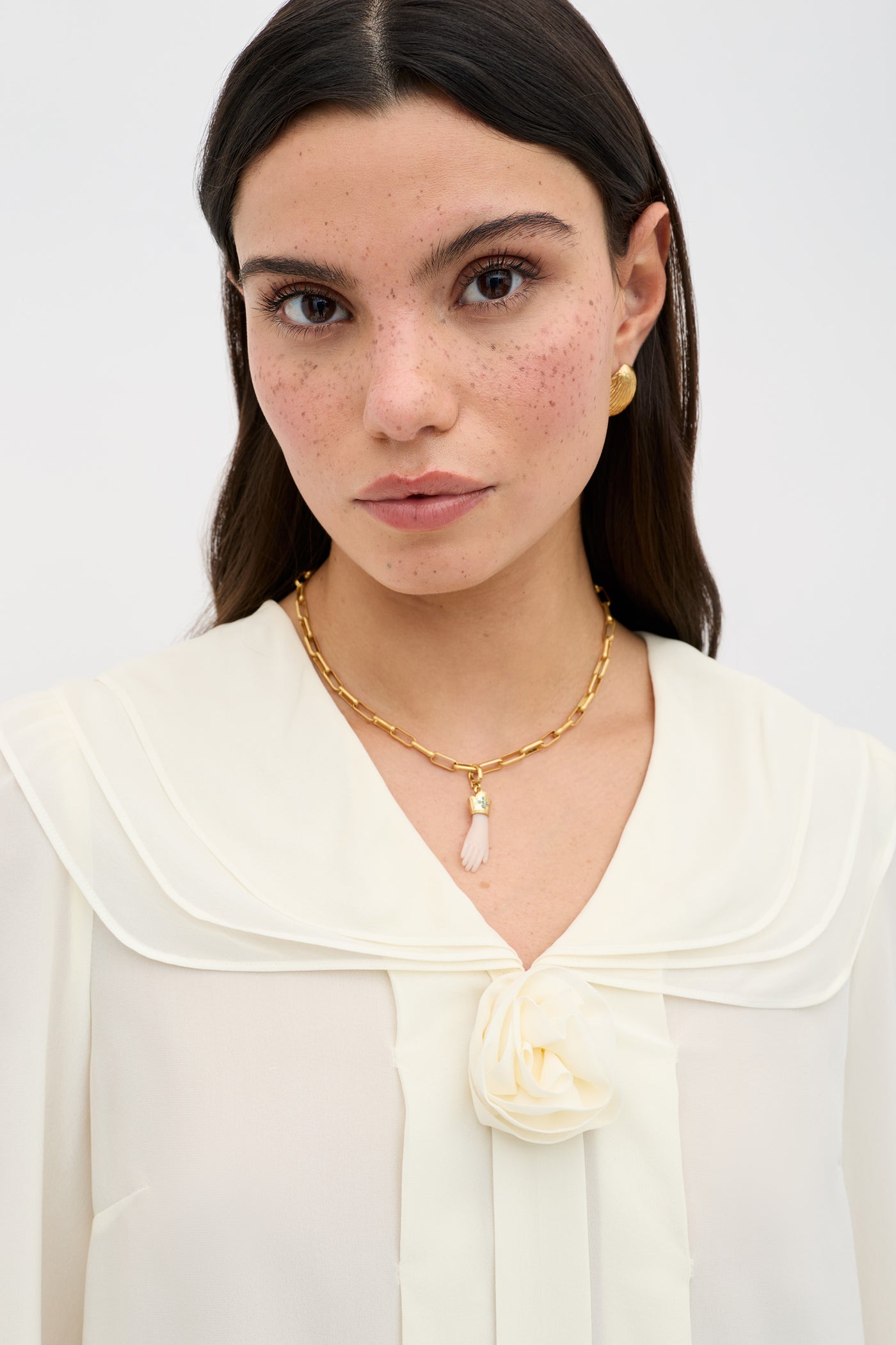 A woman with long dark hair and freckles wears the Emelin - Ivory blouse, featuring a decorative flower at the collar, paired with gold pendant necklace and earrings, standing against a plain light background.