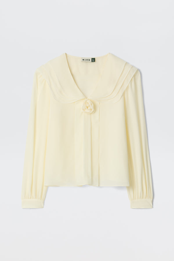 The Emelin - Ivory is a cream-colored, long-sleeve blouse featuring a wide sailor collar, fabric flower at the front, and gathered cuffs, shown against a plain white background.