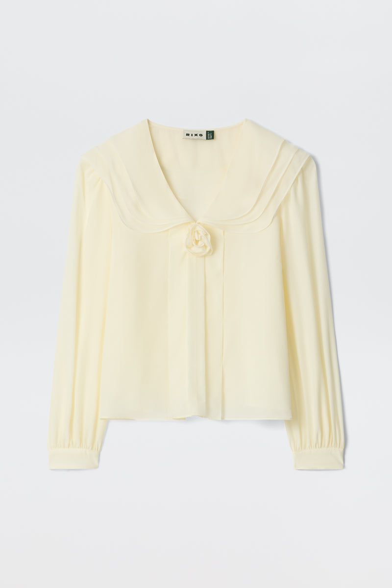 The Emelin - Ivory is a cream-colored, long-sleeve blouse featuring a wide sailor collar, fabric flower at the front, and gathered cuffs, shown against a plain white background.