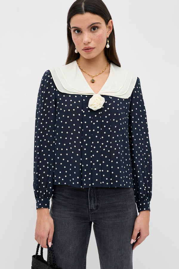 A woman in the Emelin Layla Dot Navy blouse with white polka dots, a cream sailor collar, and floral detail at the neckline, paired with dark jeans and a black textured handbag. She has long straight brown hair.