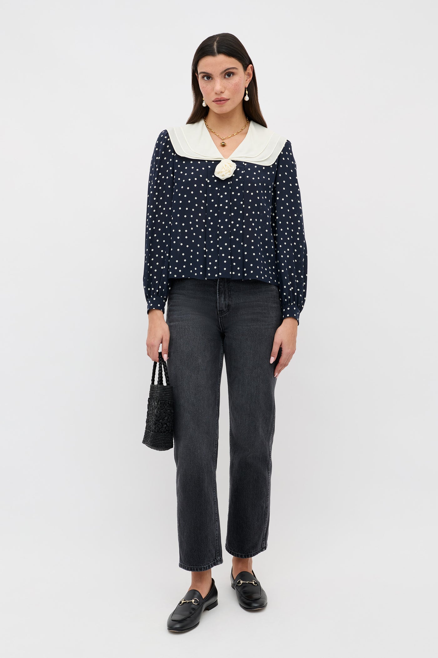 A woman stands against a plain background wearing the Emelin Layla Dot Navy blouse with white polka dots and a large white collar, black straight-leg jeans, black loafers, and holding a small black textured handbag.
