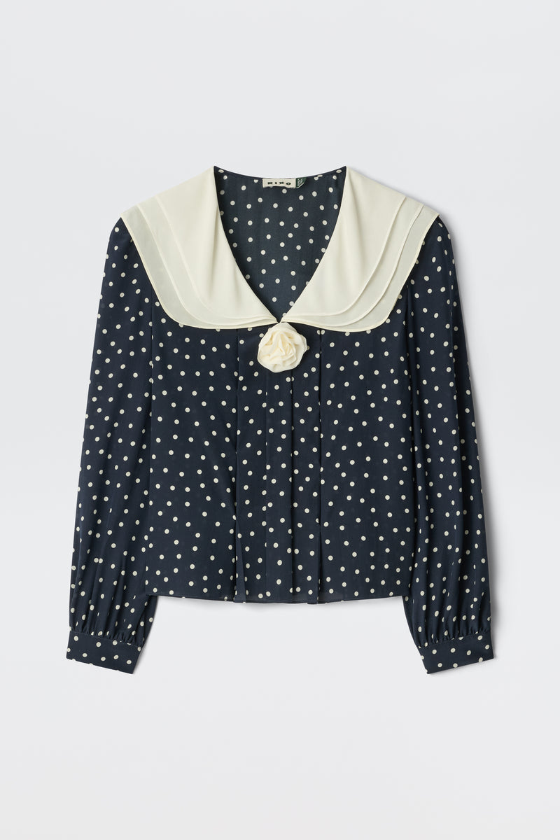 The Emelin Layla Dot Navy is a navy blue blouse with white polka dots, long sleeves, a wide cream sailor collar, and a decorative cream fabric flower at the front.