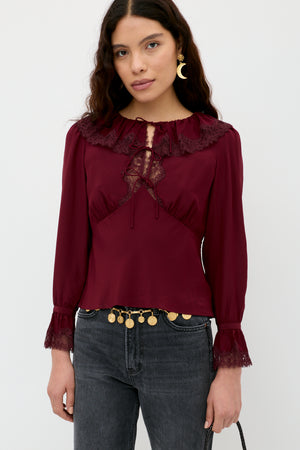 A woman with long wavy hair wears the Aimme - Maroon blouse, featuring lace details, long sleeves, and a ruffled collar, paired with dark gray jeans and a gold coin belt. She stands against a plain white background.