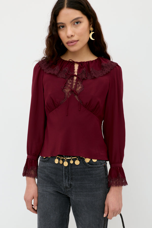 A woman with long wavy hair wears the Aimme - Maroon blouse, featuring lace details, long sleeves, and a ruffled collar, paired with dark gray jeans and a gold coin belt. She stands against a plain white background.
