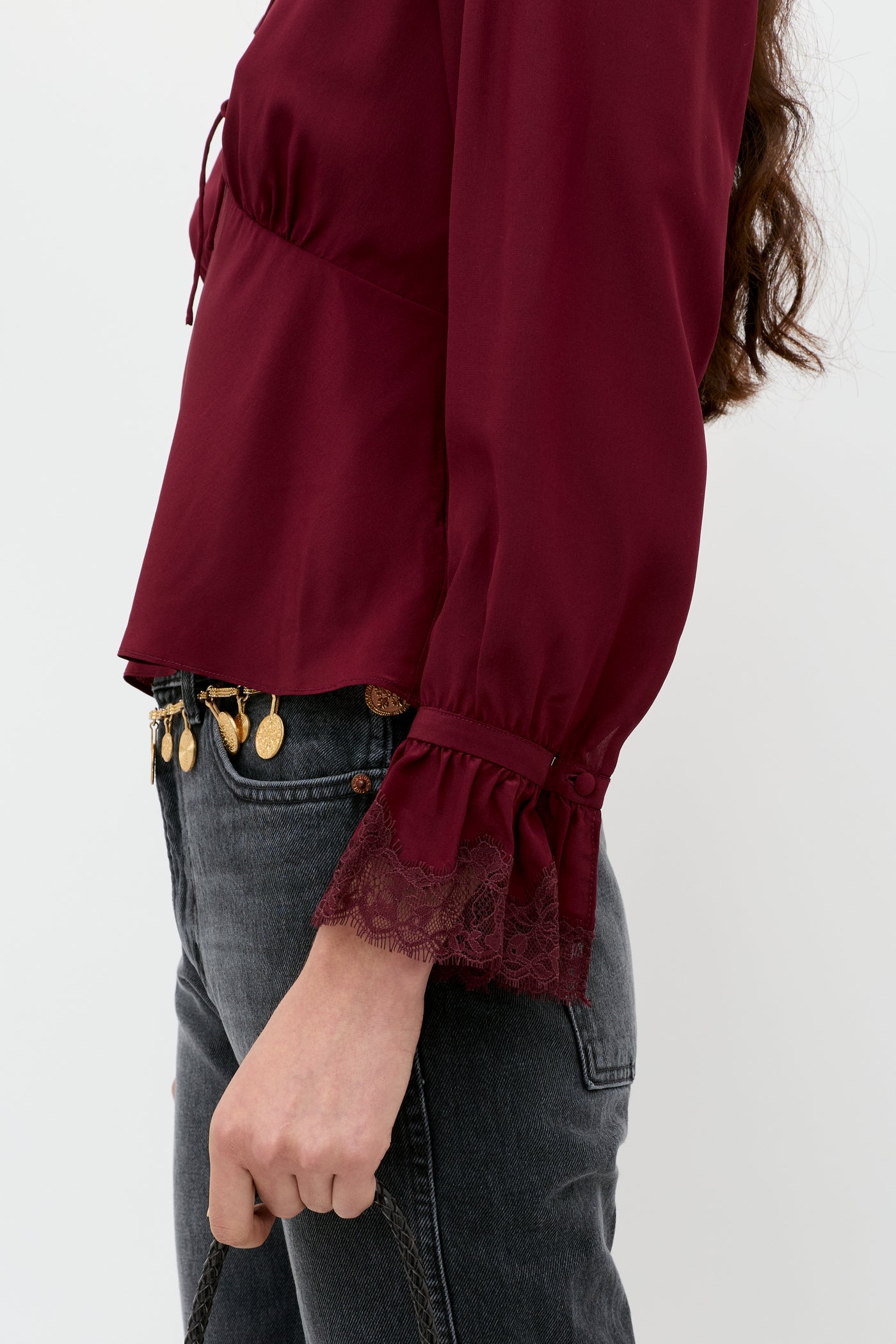 A person wears the Aimme – Maroon long-sleeved blouse with lace cuffs and dark jeans, holding a black strap. Gold coin accessories accent the waist against a plain white background.
