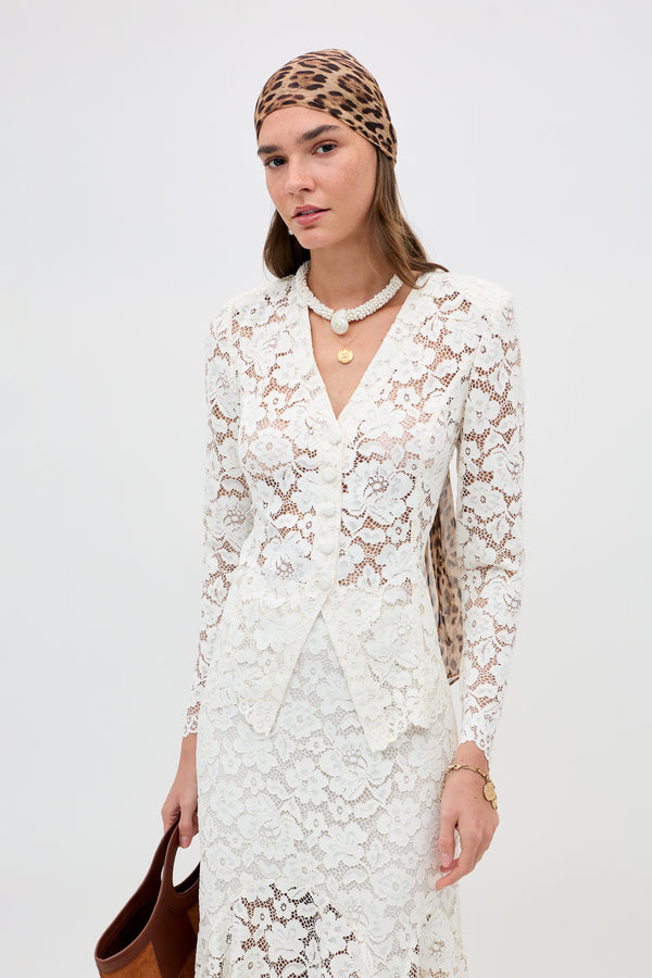 A woman in the Padma - Cream long-sleeve lace dress, leopard print headscarf, pearl pendant necklace, and holding a brown bag stands against a plain white background.