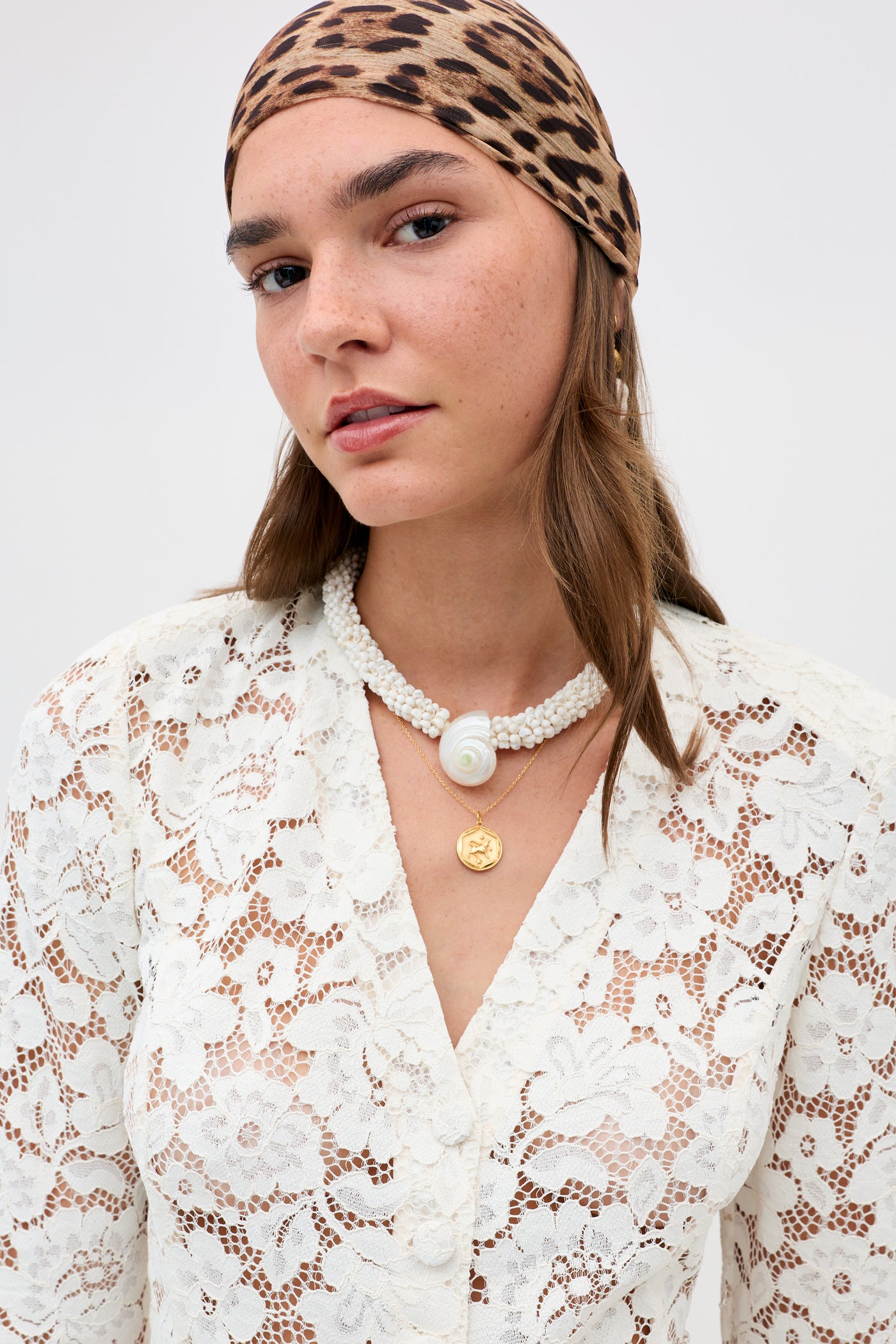 A woman wearing the Padma - Cream leopard-print headscarf, a white lace blouse, a shell pendant necklace, and a gold pendant necklace looks at the camera against a plain background.