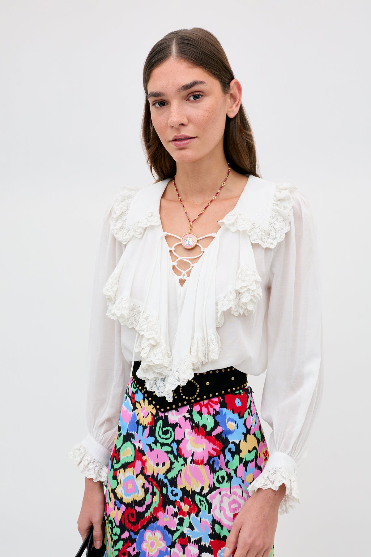A woman with long brown hair wears the Milia - White blouse with lace and ruffle details, paired with a floral skirt. She accessorizes with a cameo necklace and black bag, standing against a plain light background.