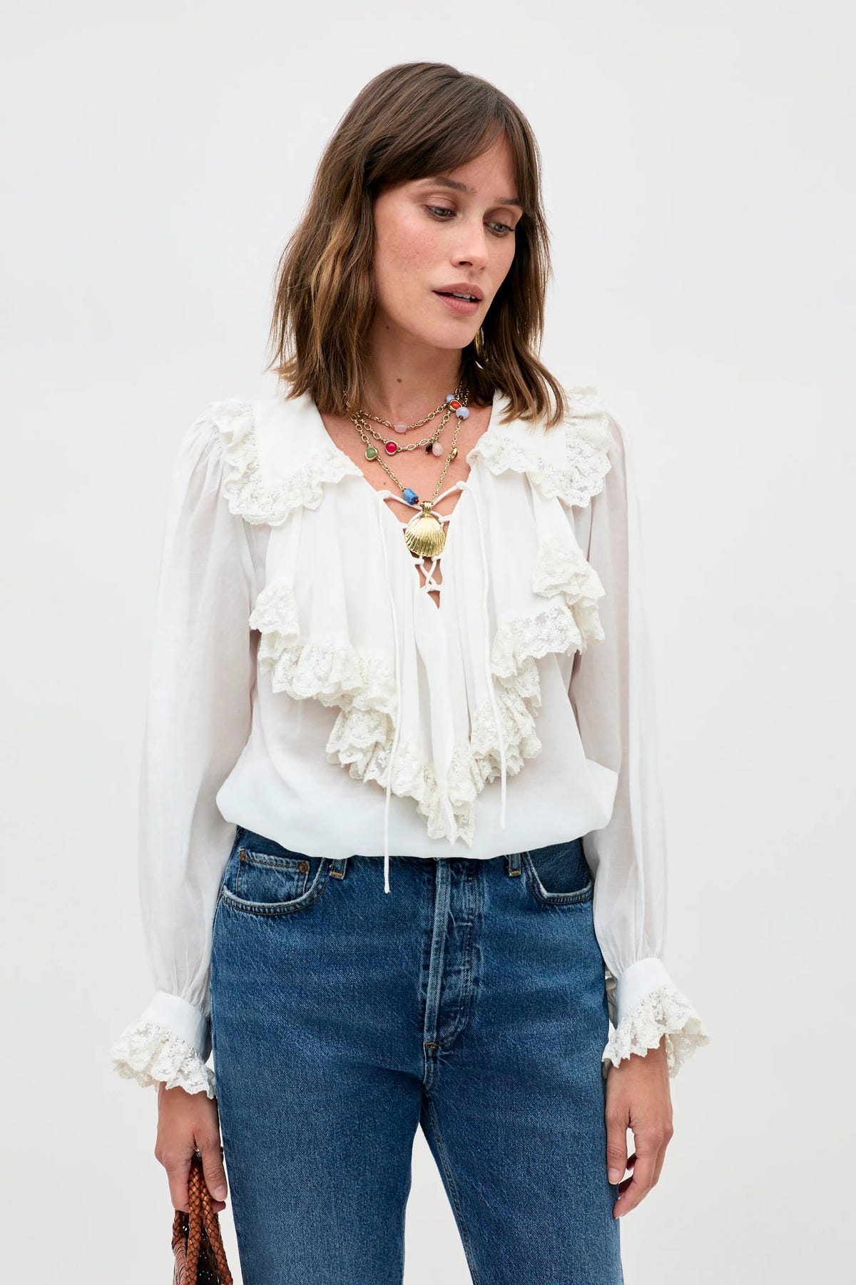 A woman wears the Milia - White blouse, featuring long sleeves and ruffled lace, paired with blue jeans and layered colorful necklaces, standing against a light background while holding a brown item in her left hand.