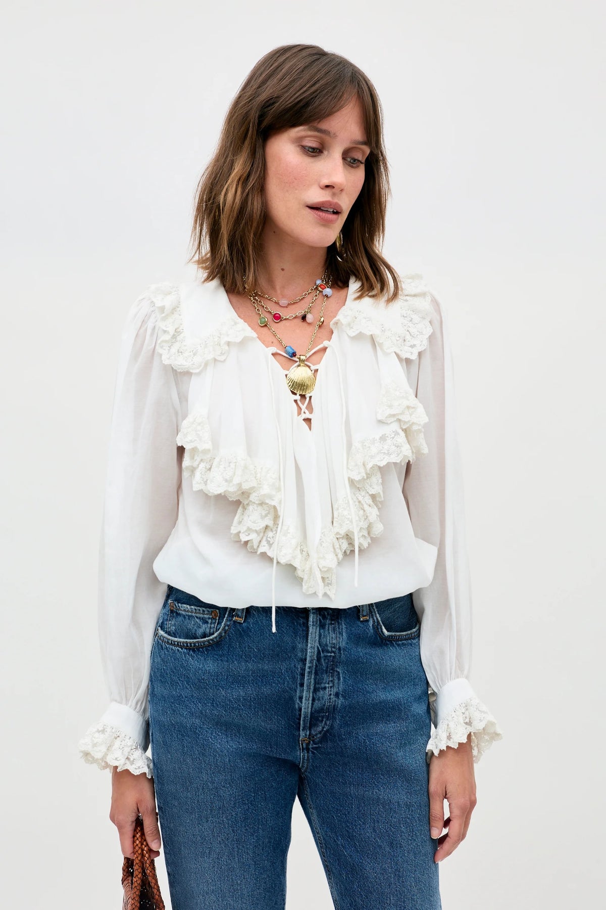 A woman wears the Milia - White blouse, featuring long sleeves and ruffled lace, paired with blue jeans and layered colorful necklaces, standing against a light background while holding a brown item in her left hand.