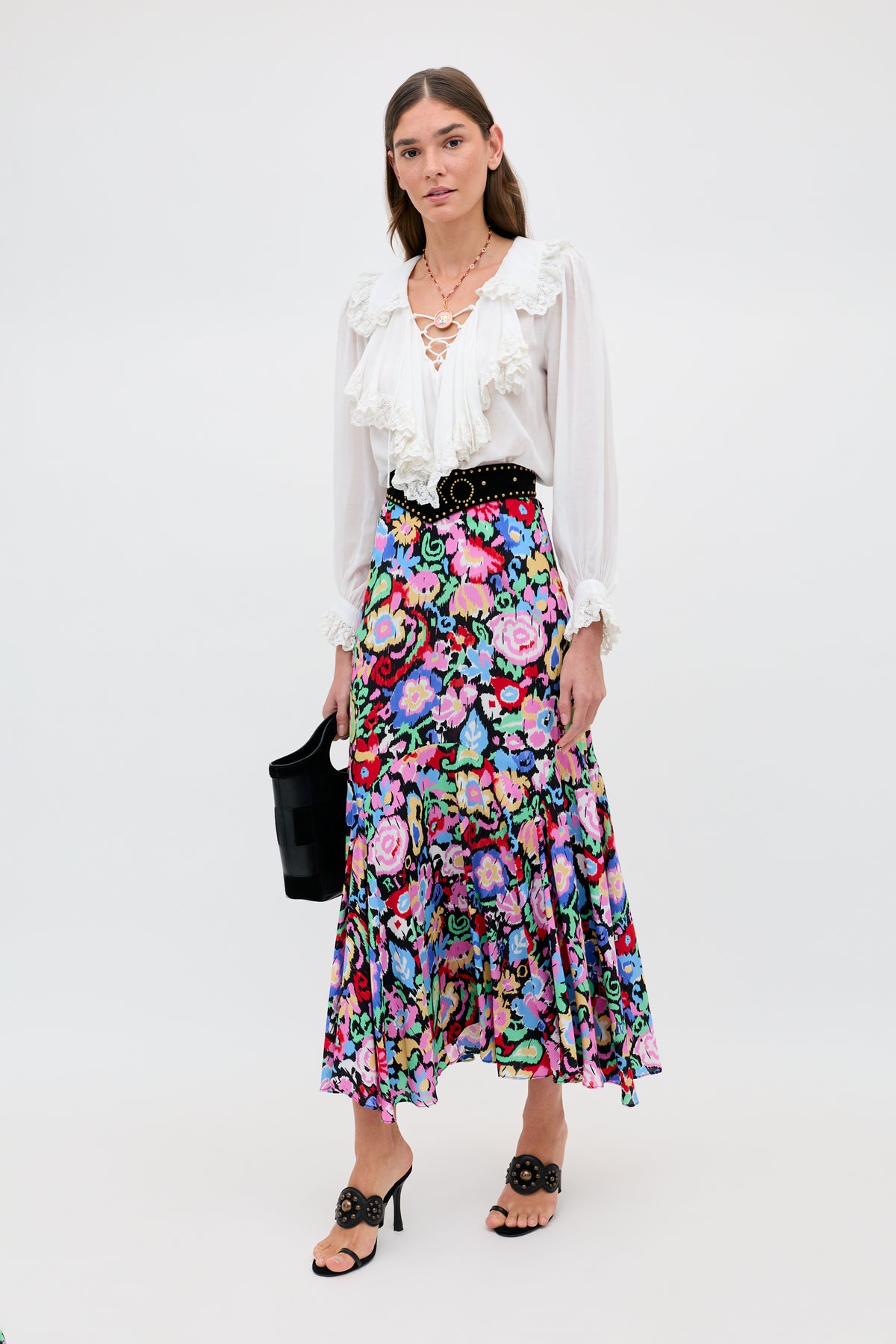 A woman with long brown hair and a calm expression stands against a plain background wearing the Milia - White ruffled blouse, a colorful floral midi skirt, black sandals, and holding a black handbag.