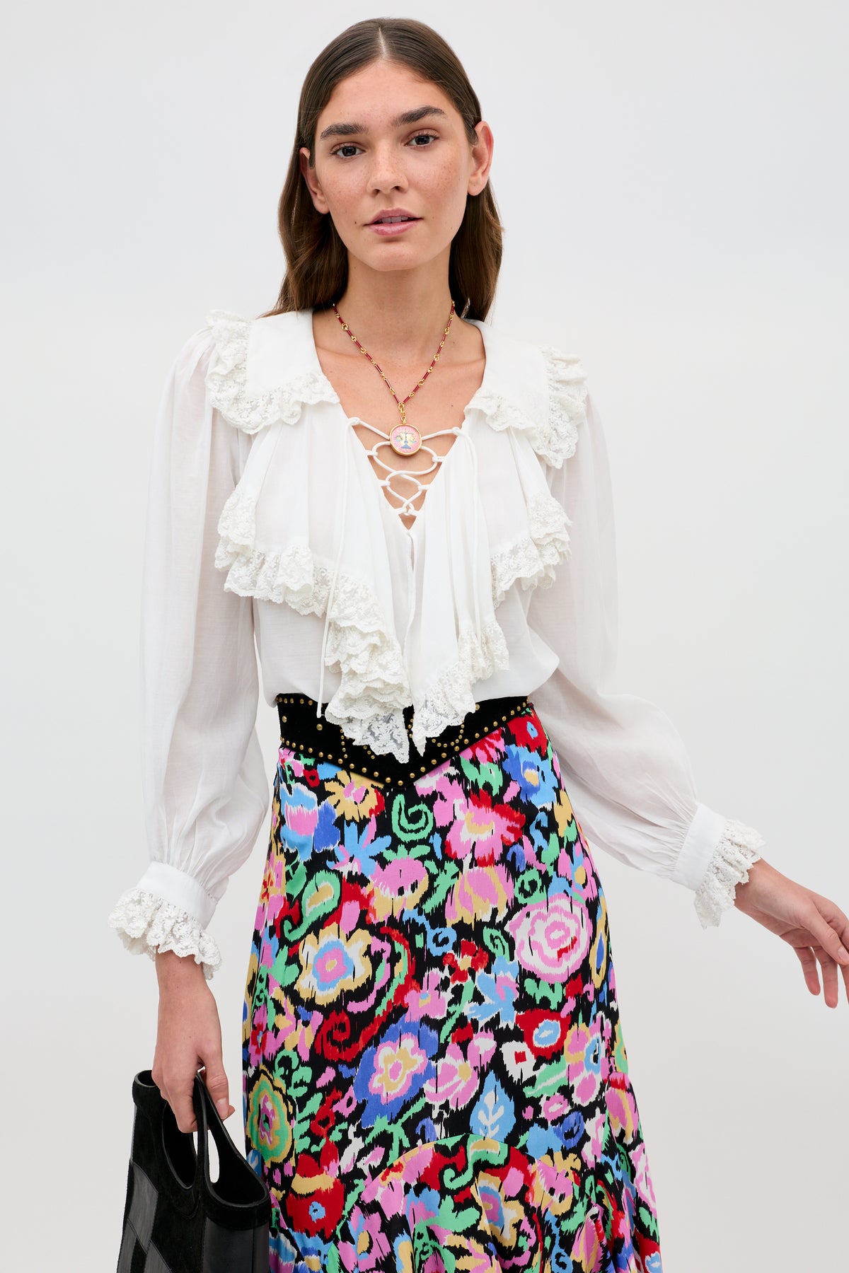 A woman wears the Milia - White blouse, featuring ruffled lace details, paired with a colorful floral skirt and holding a black handbag against a plain background.