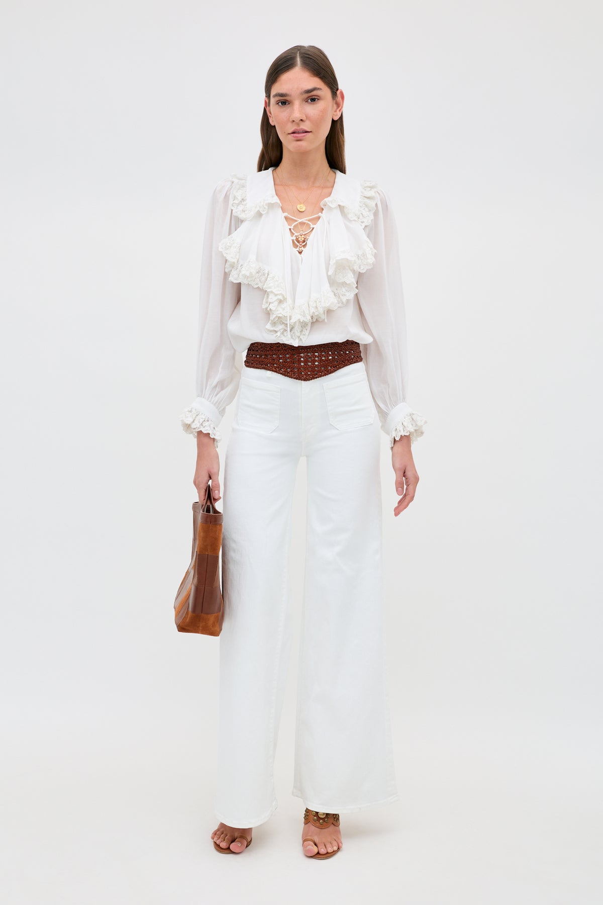 A woman stands against a white background wearing the Milia - White blouse paired with high-waisted white pants, a brown braided belt, brown heeled sandals, and holding a brown handbag.