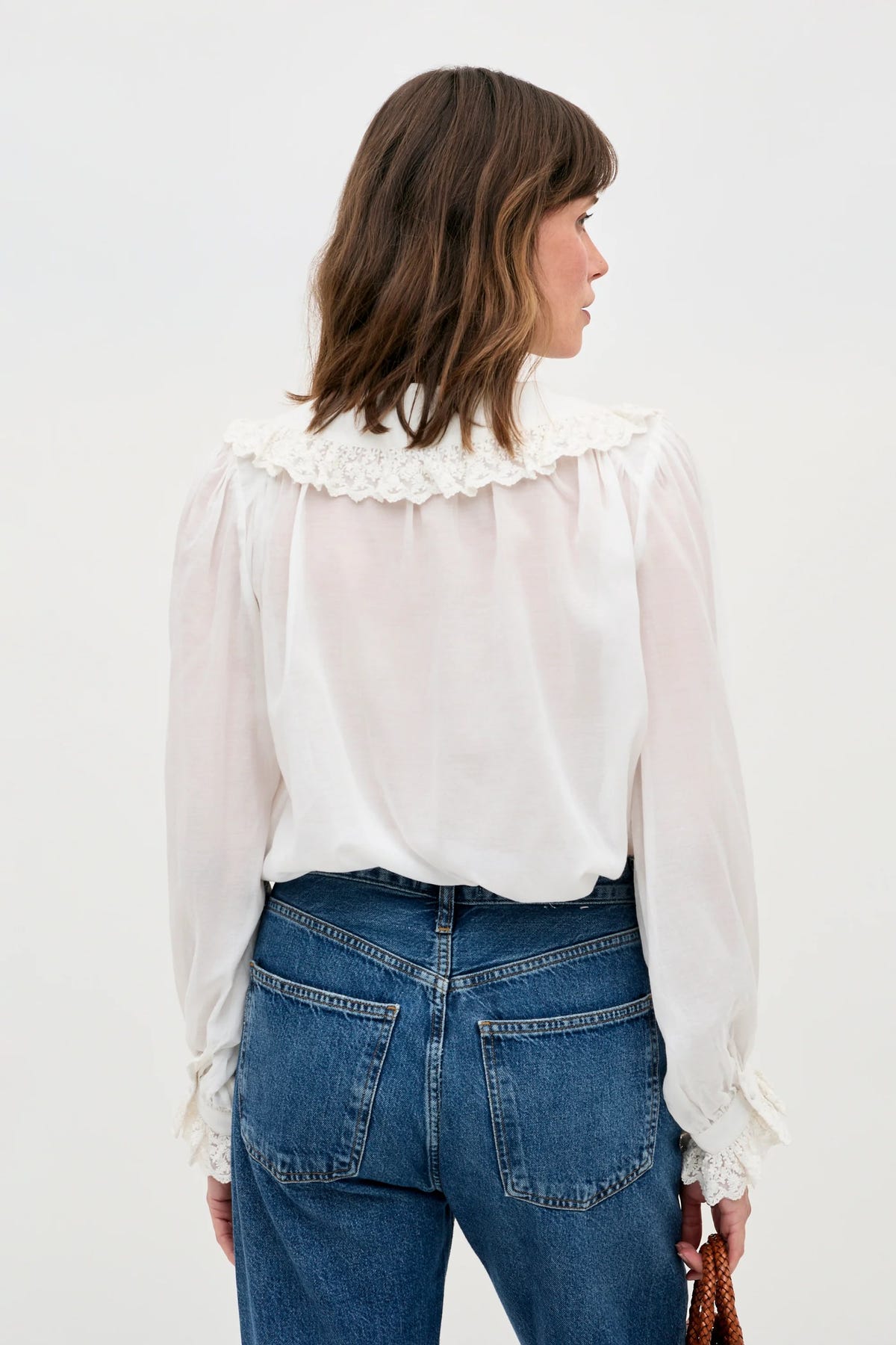 A woman with shoulder-length brown hair, seen from behind, wears the Milia - White blouse, featuring lace details on the collar and cuffs, paired with high-waisted blue jeans. She carries a brown woven handbag.
