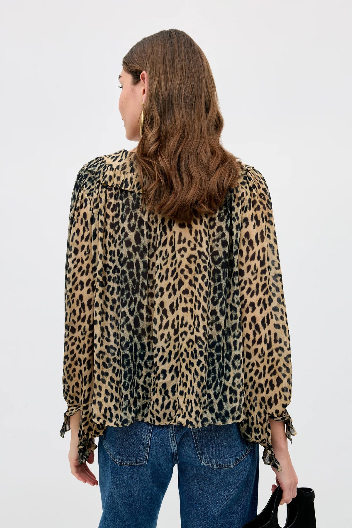 A woman with wavy brown hair, seen from behind, wears the Milia - Nina Leopard Brown blouse—a loose, long-sleeve leopard print top—paired with blue jeans and holding a black bag. The background is plain white.