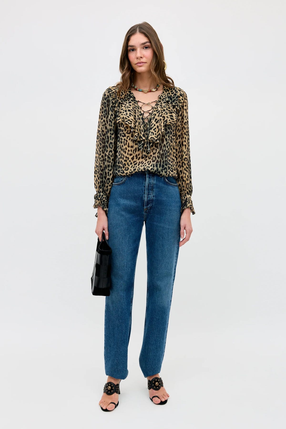 A woman stands against a white background wearing the Milia - Nina Leopard Brown blouse, paired with blue high-waisted jeans, black heeled sandals, a chunky necklace, and holding a small black handbag.