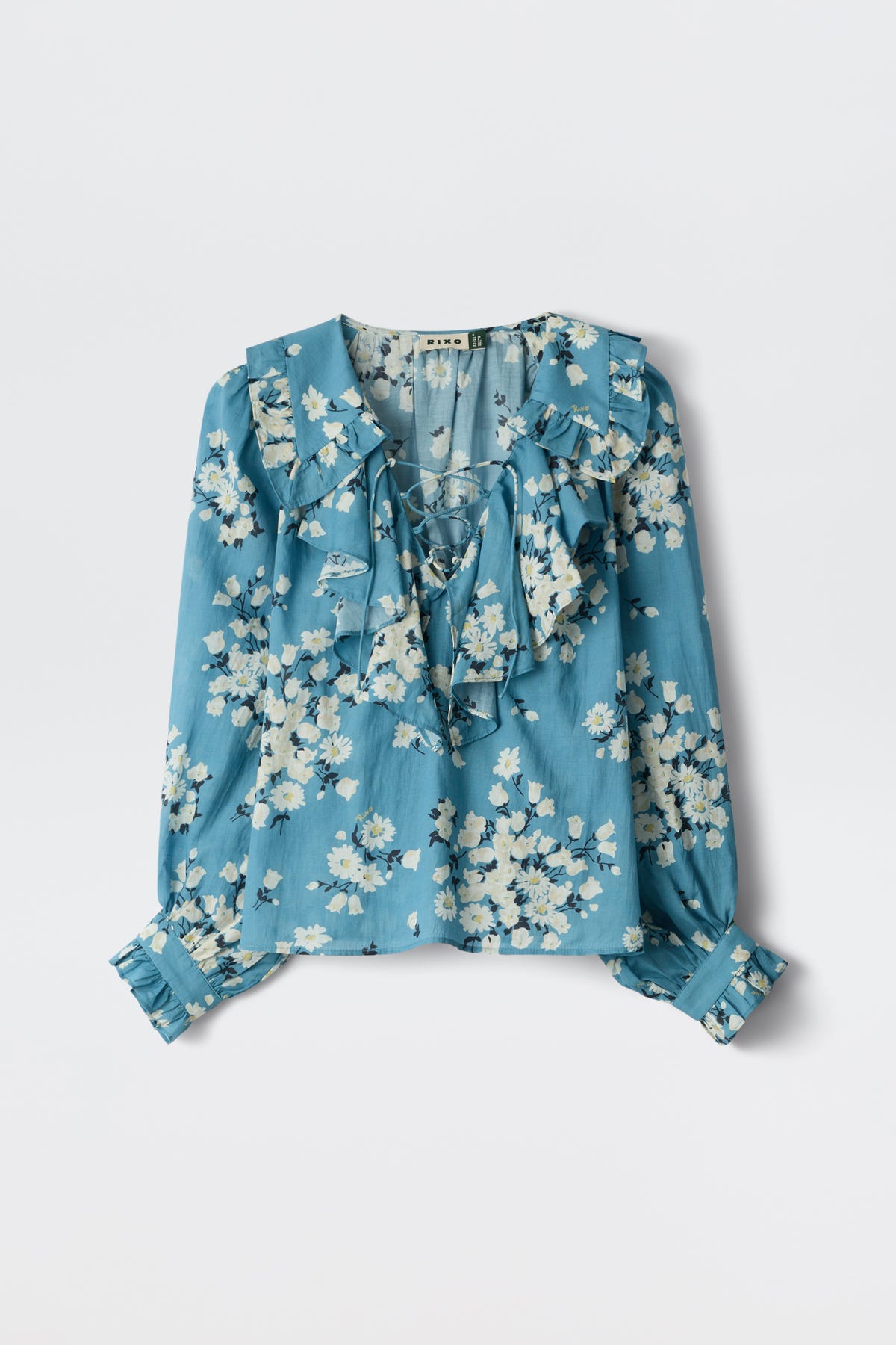 The Milia - Amelia Bouquet Blue is a long-sleeve blue blouse featuring a white floral print, ruffled collar and front, buttoned cuffs, and a loose, flowy fit.