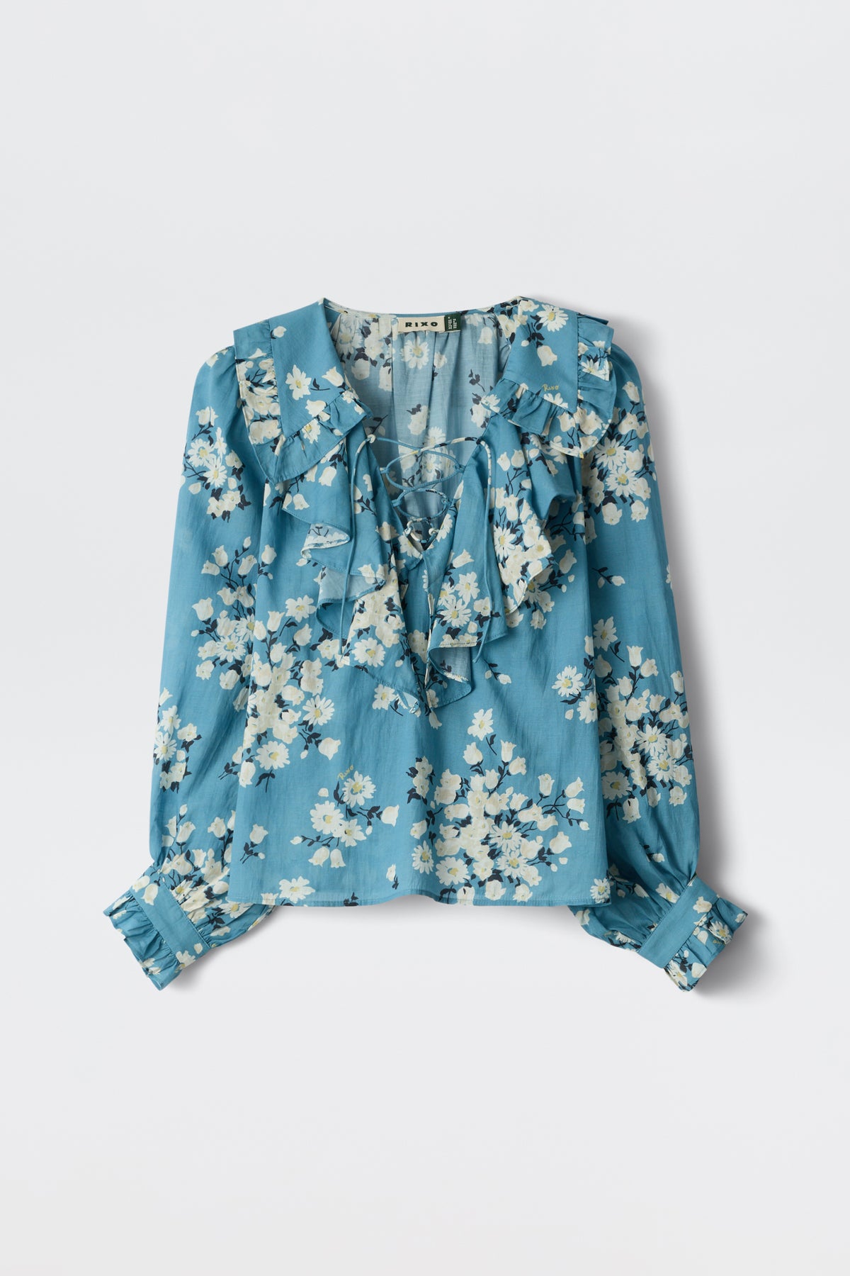 The Milia - Amelia Bouquet Blue is a long-sleeve blue blouse featuring a white floral print, ruffled collar and front, buttoned cuffs, and a loose, flowy fit.