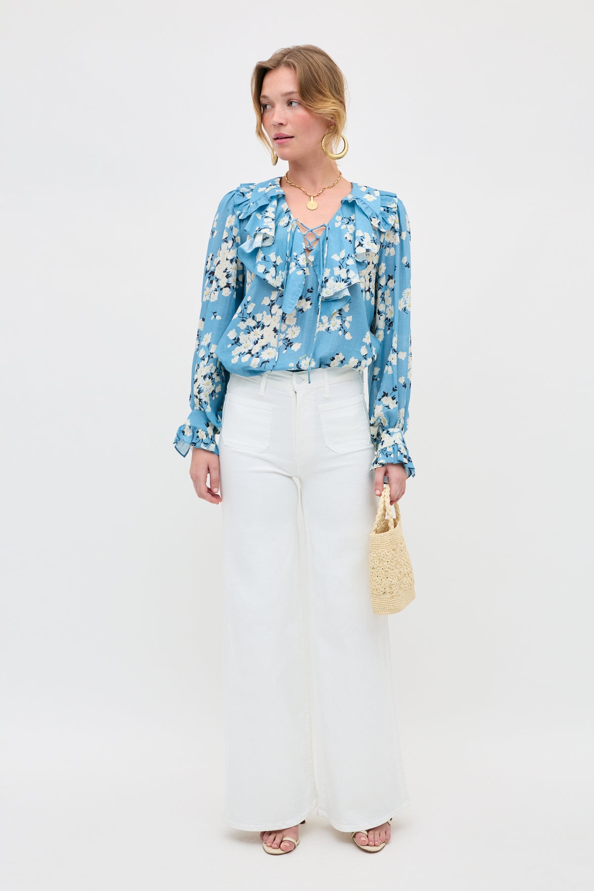 A woman stands against a white backdrop wearing the Milia - Amelia Bouquet Blue floral blouse with ruffles, high-waisted white wide-leg pants, sandals, a woven handbag, gold hoop earrings, and layered necklaces.