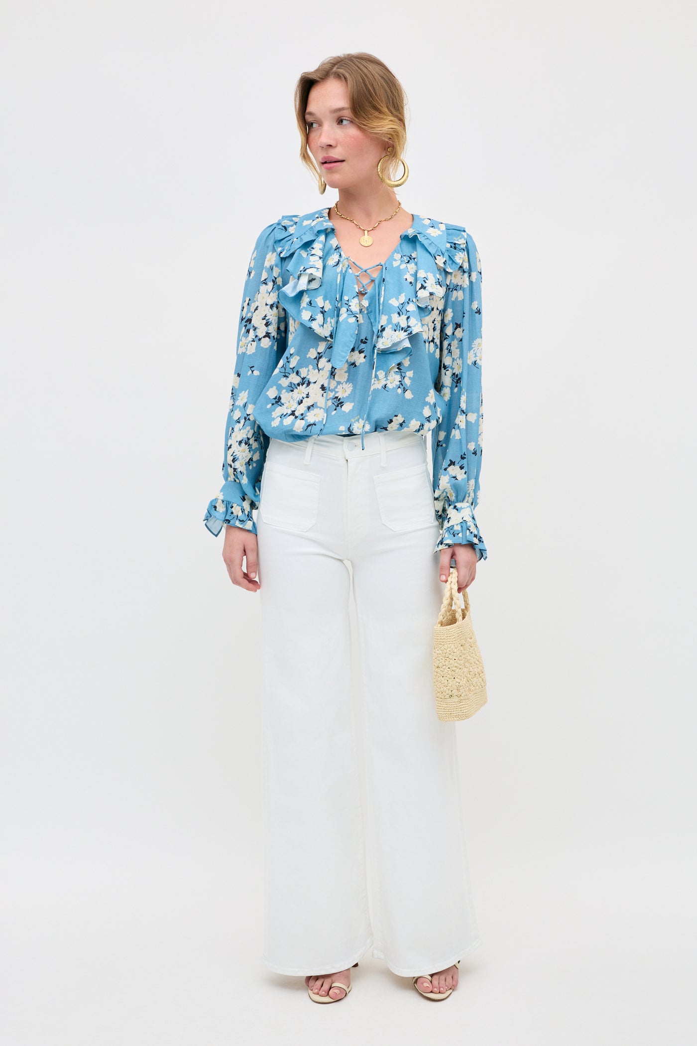 A woman stands against a white backdrop wearing the Milia - Amelia Bouquet Blue floral blouse with ruffles, high-waisted white wide-leg pants, sandals, a woven handbag, gold hoop earrings, and layered necklaces.