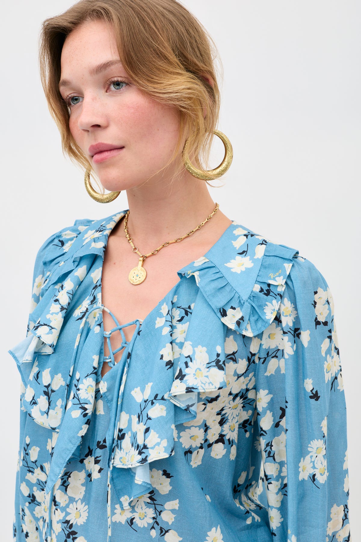 A woman with light skin and blonde hair in a loose updo wears a blue floral ruffle blouse, large gold hoop earrings, and the Sol - Gold pendant on an adjustable chain, posing against a plain light background.