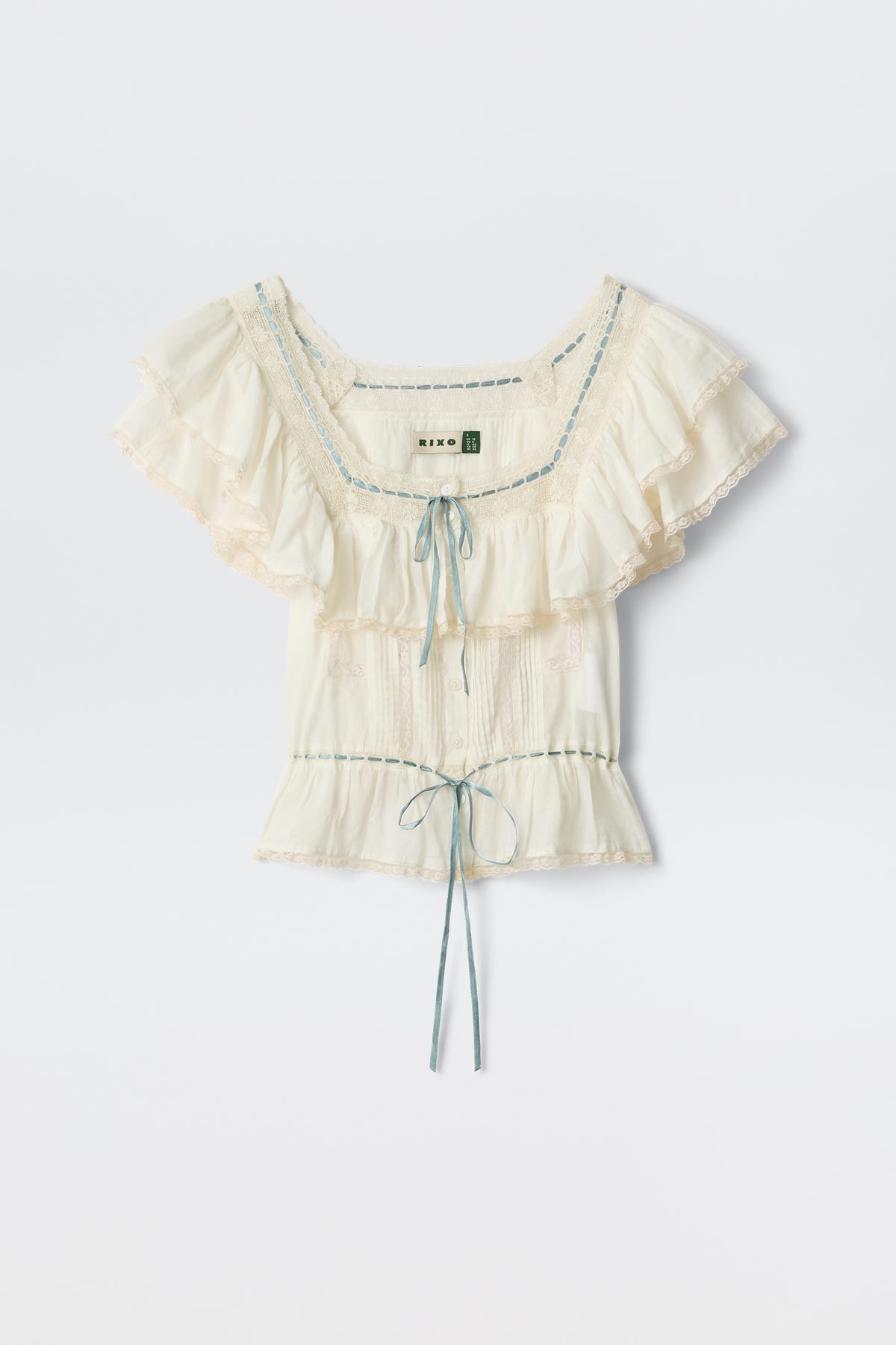 The Melisa - Ivory blouse features a prairie-inspired design with cream ruffles, lace trim, blue ribbon details, a gathered bust, and a waist tie, shown on a plain white background.