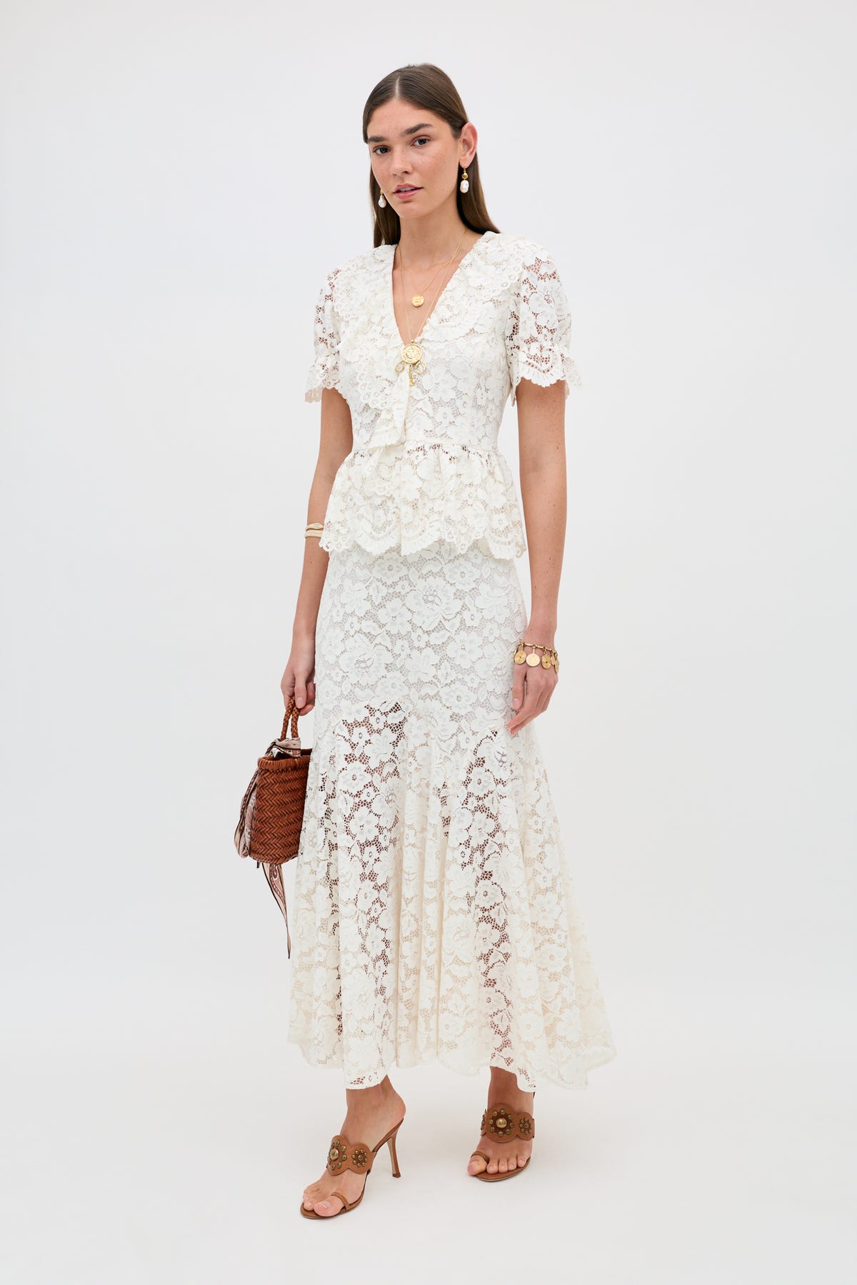 A woman stands against a white background wearing the Dollie - Cream lace short-sleeve peplum top with a matching long skirt, tan heeled sandals, gold jewelry, and holding a brown woven handbag.