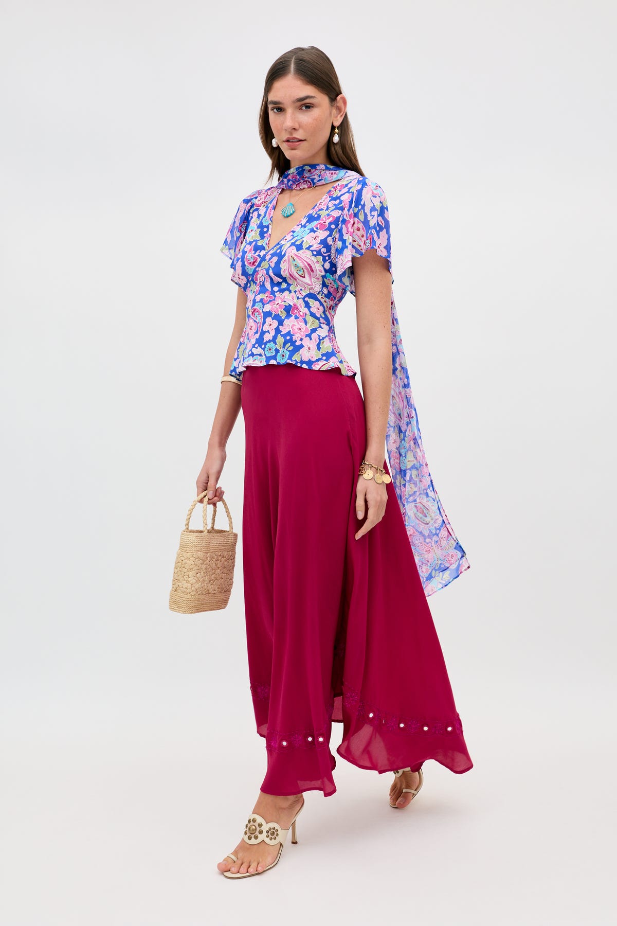 A woman wearing a blue floral top with sheer cape, beige heels, hoop earrings, and holding a woven handbag pairs her outfit with the Rudy - Mirror Embellishment Magenta Pink flowy pants as she poses against a plain white background.