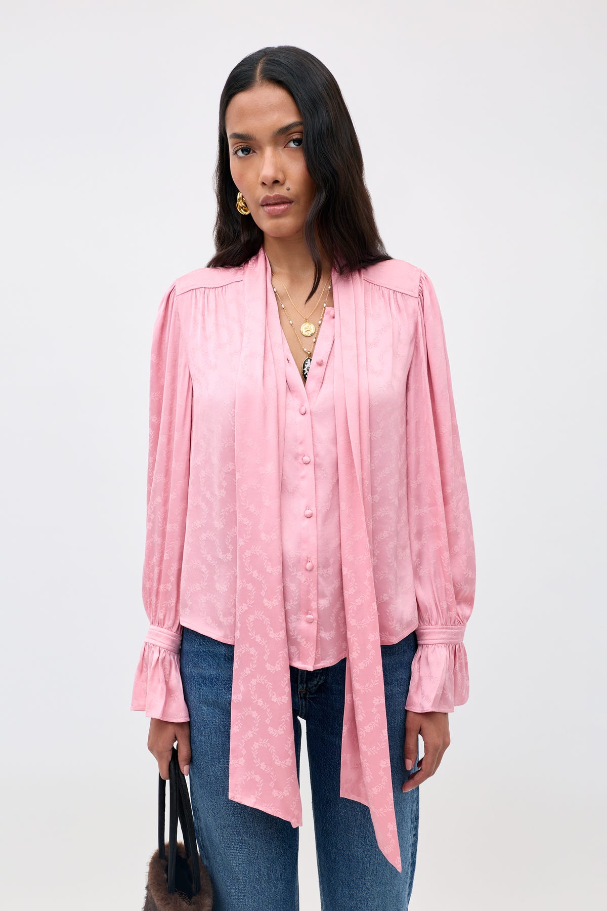 A woman wearing the Evanora - Floral Trail pink, long-sleeved blouse with a tie-neck detail and blue jeans stands against a plain light background, holding a handbag.