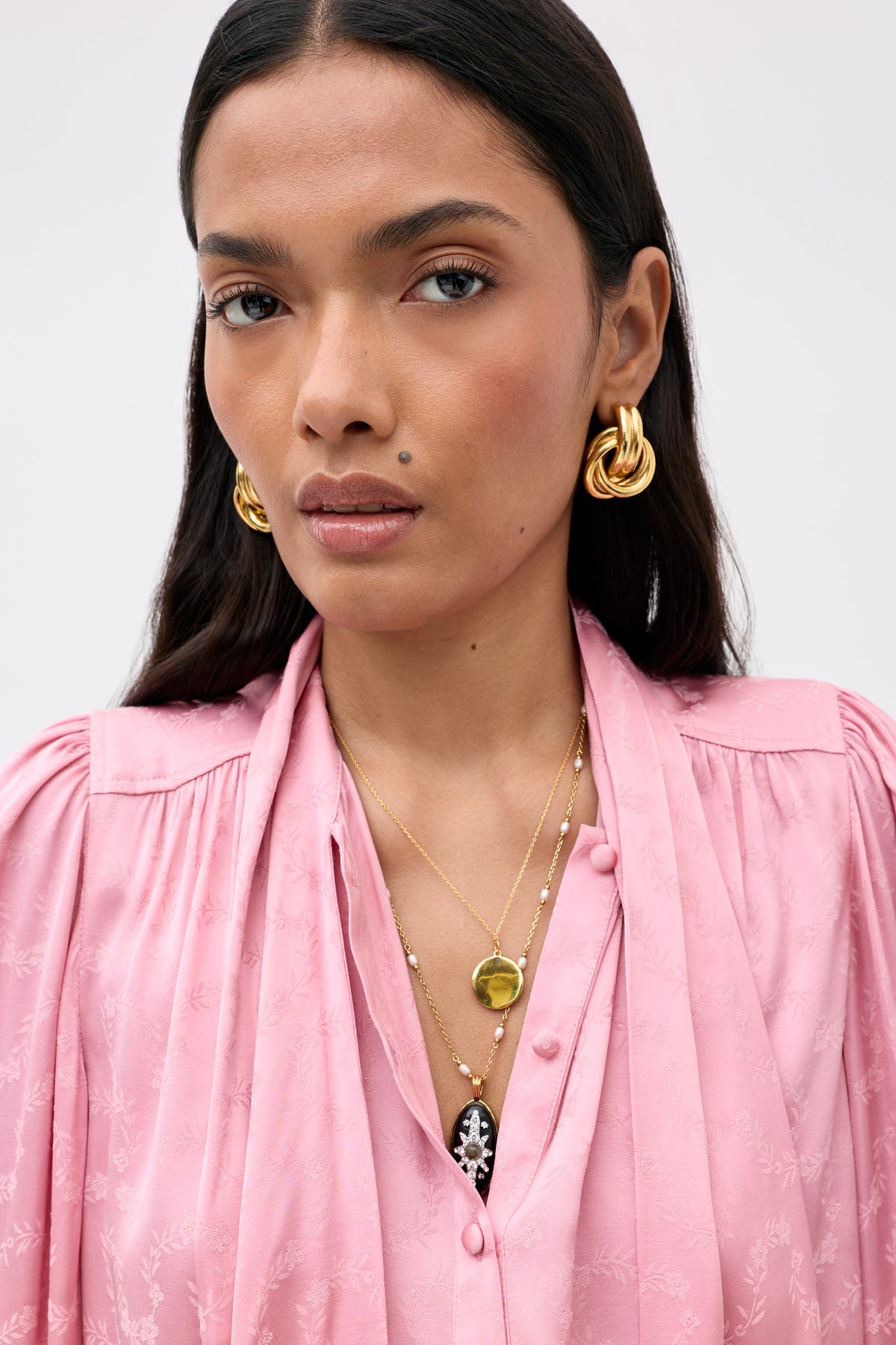 A woman in a pink blouse wears the Evanora - Floral Trail earrings, featuring gold hoops and layered gold necklaces—one adorned with a coin pendant and another with a striking black and white accent. She has long dark hair, light makeup, and a neutral expression.