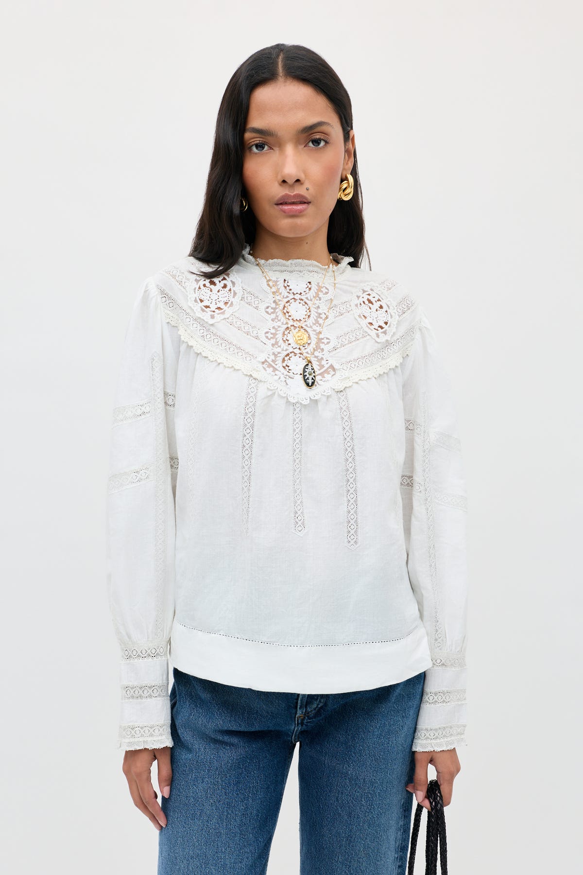 A person with long dark hair wears the Tamzin - Cream blouse with intricate lace detailing, styled with blue jeans and gold earrings, standing against a plain light background while holding a black handbag.