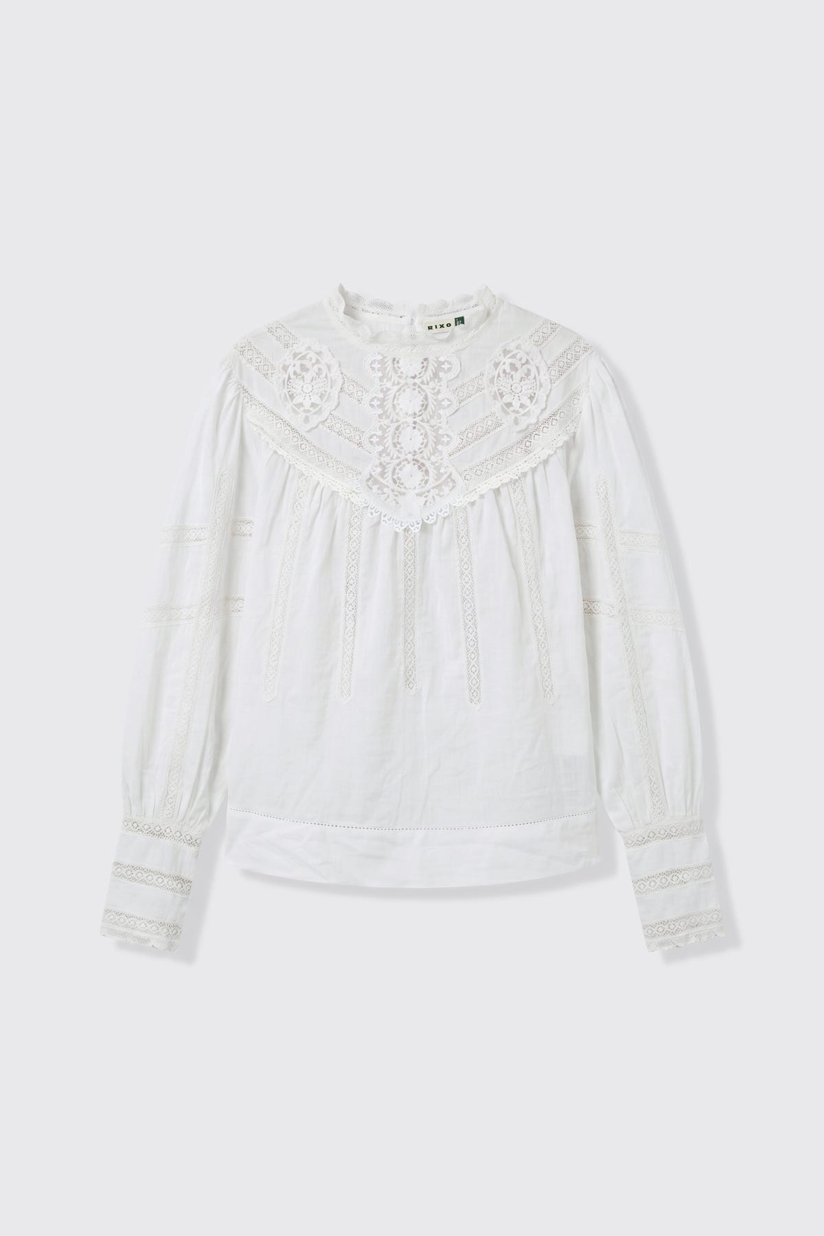 The Tamzin - Cream blouse features long sleeves and a high neckline, accentuated by lace details on the chest, sleeves, and cuffs. Its lightweight, textured fabric and intricate embroidery lend a vintage and elegant touch to the design.