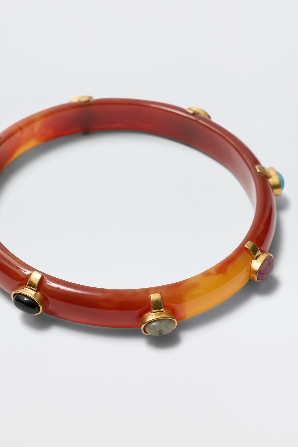 The Sabella - Tortoise is a reddish-brown marbled resin bangle with multicoloured cabochons in gold-plated bezels spaced around its circumference, shown on a white background.