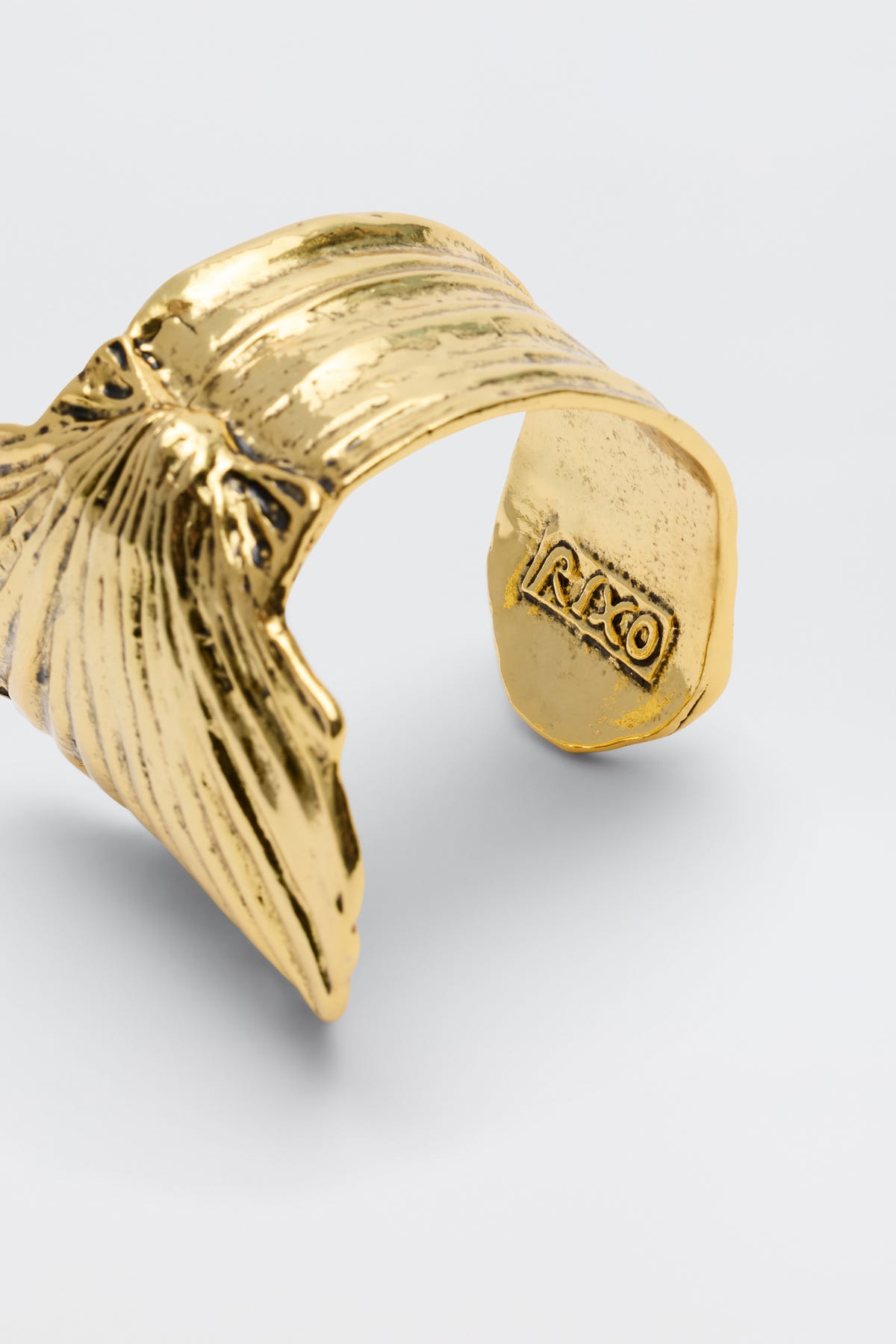 The Salene - Gold ring, crafted from gold-plated brass in the form of a sculpted leaf or wing with intricate grooves and the word "RAXO" engraved on its inner band, is displayed against a simple light gray background. Its antique finish and elegant design evoke the classic beauty of shell-inspired jewelry.