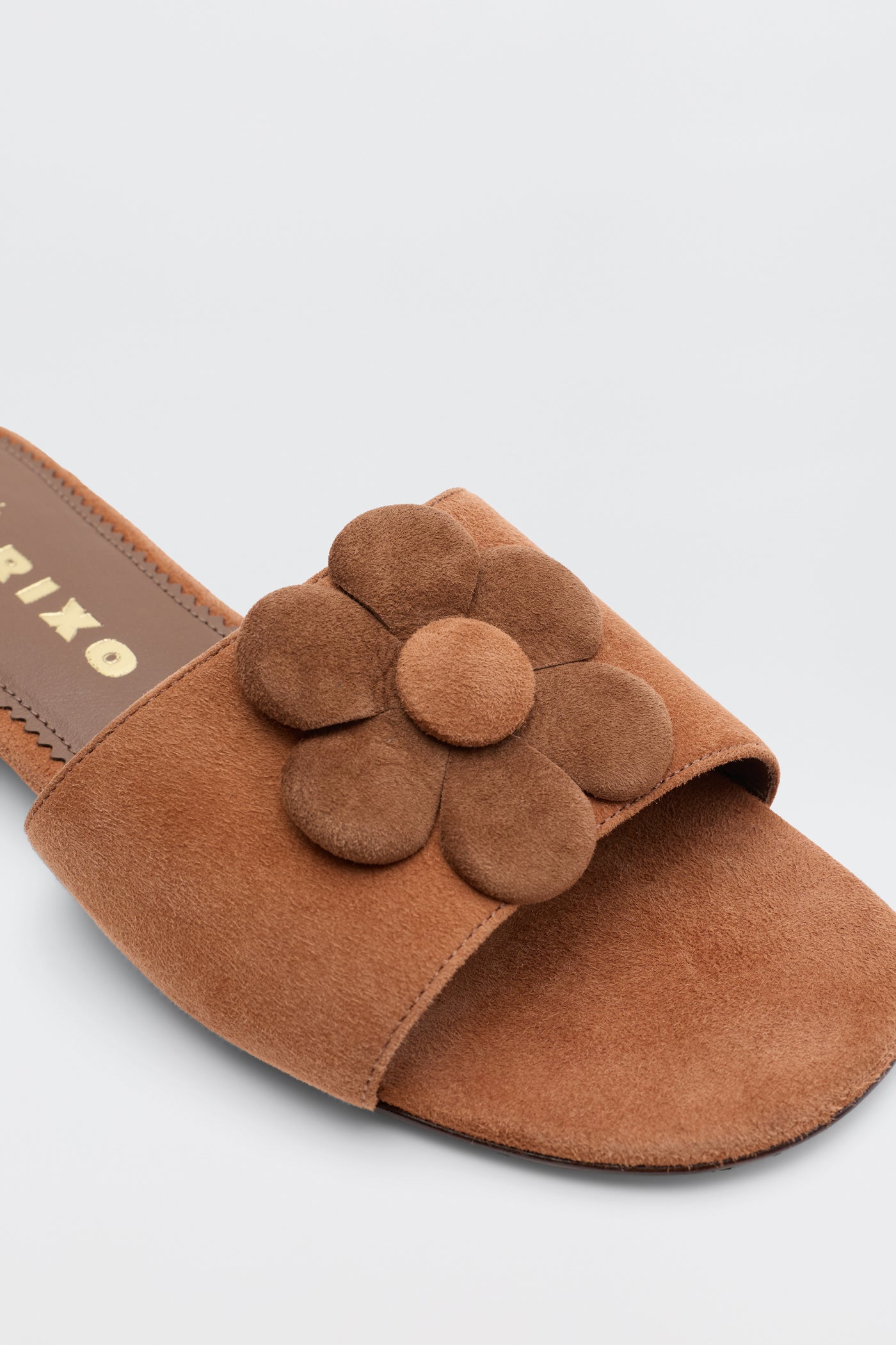 The Kenna - Brown sandal features brown suede with a wide strap and matching suede flower, set against a plain light background. Perfect for summer looks, its insole is finished with elegant gold lettering.