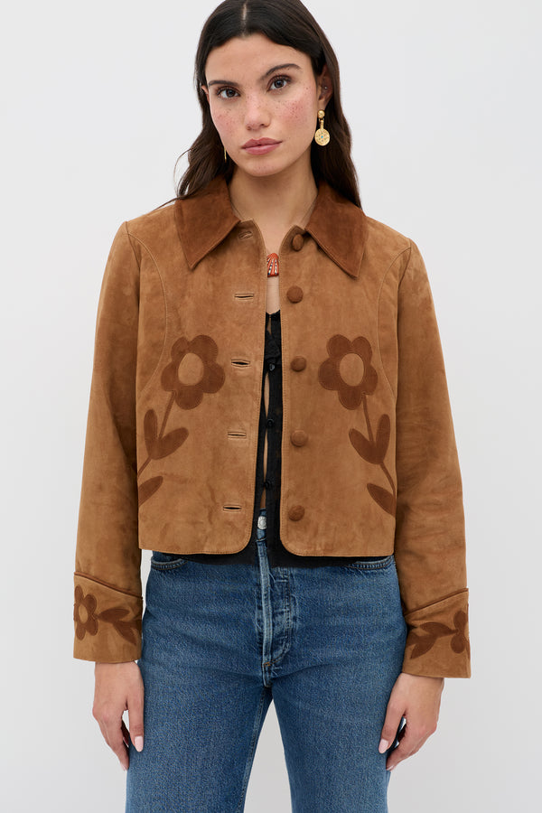 A woman wears the Dionne - Retro Daisy tan suede jacket with large daisy appliqué on the front and cuffs, paired with blue jeans, standing against a plain white background.