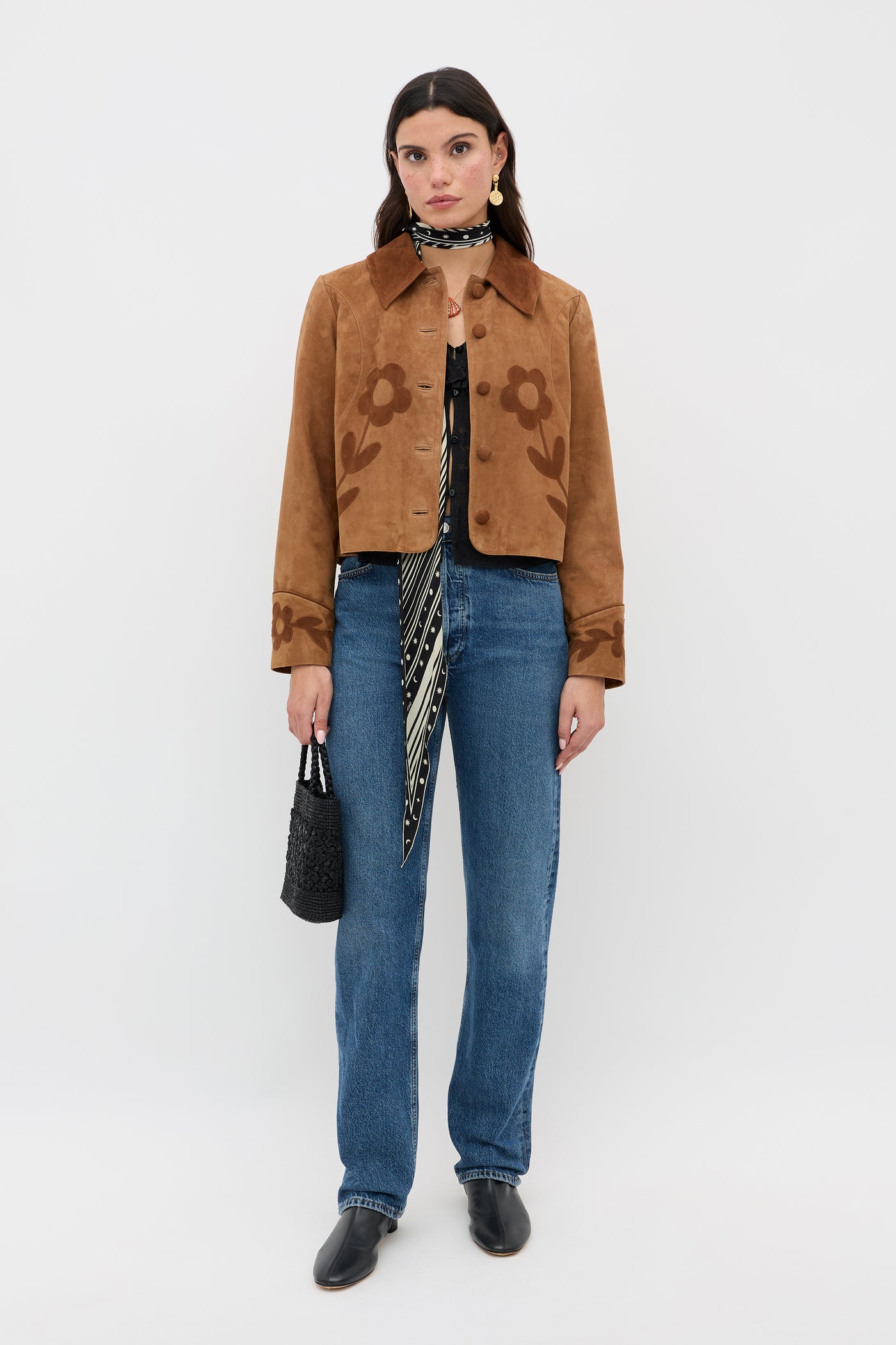 A woman poses against a plain backdrop wearing the Dionne - Retro Daisy brown suede jacket with daisy appliqué, a patterned scarf, blue jeans, black shoes, and carrying a small black textured handbag.