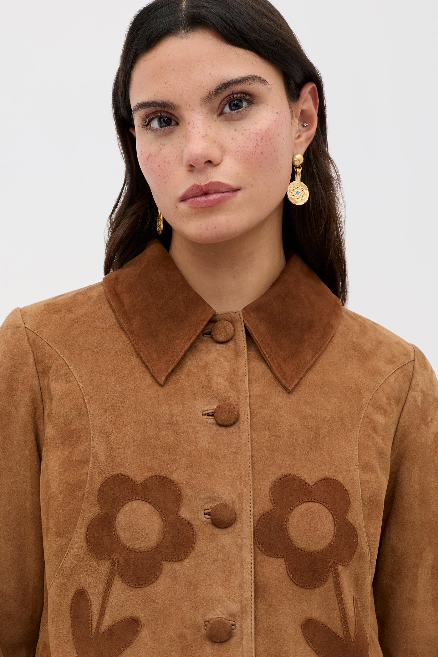 A woman with long dark hair and freckles wears the Dionne - Retro Daisy tan suede jacket with a large collar and daisy appliqué. She has gold drop earrings, a neutral expression, and stands against a plain light background.