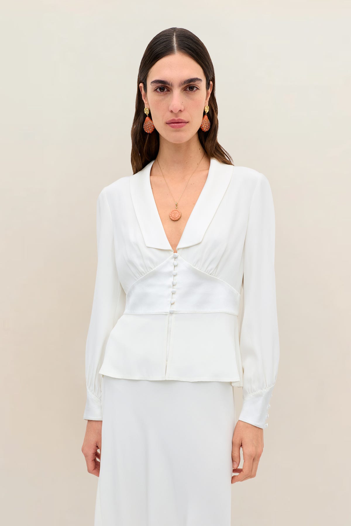 A woman stands against a plain beige background, wearing the Adeline - Ivory silk-crepe long-sleeve blouse featuring a deep V-neck and decorative buttons reminiscent of elegant bridal tops, paired with a pendant necklace and orange drop earrings.