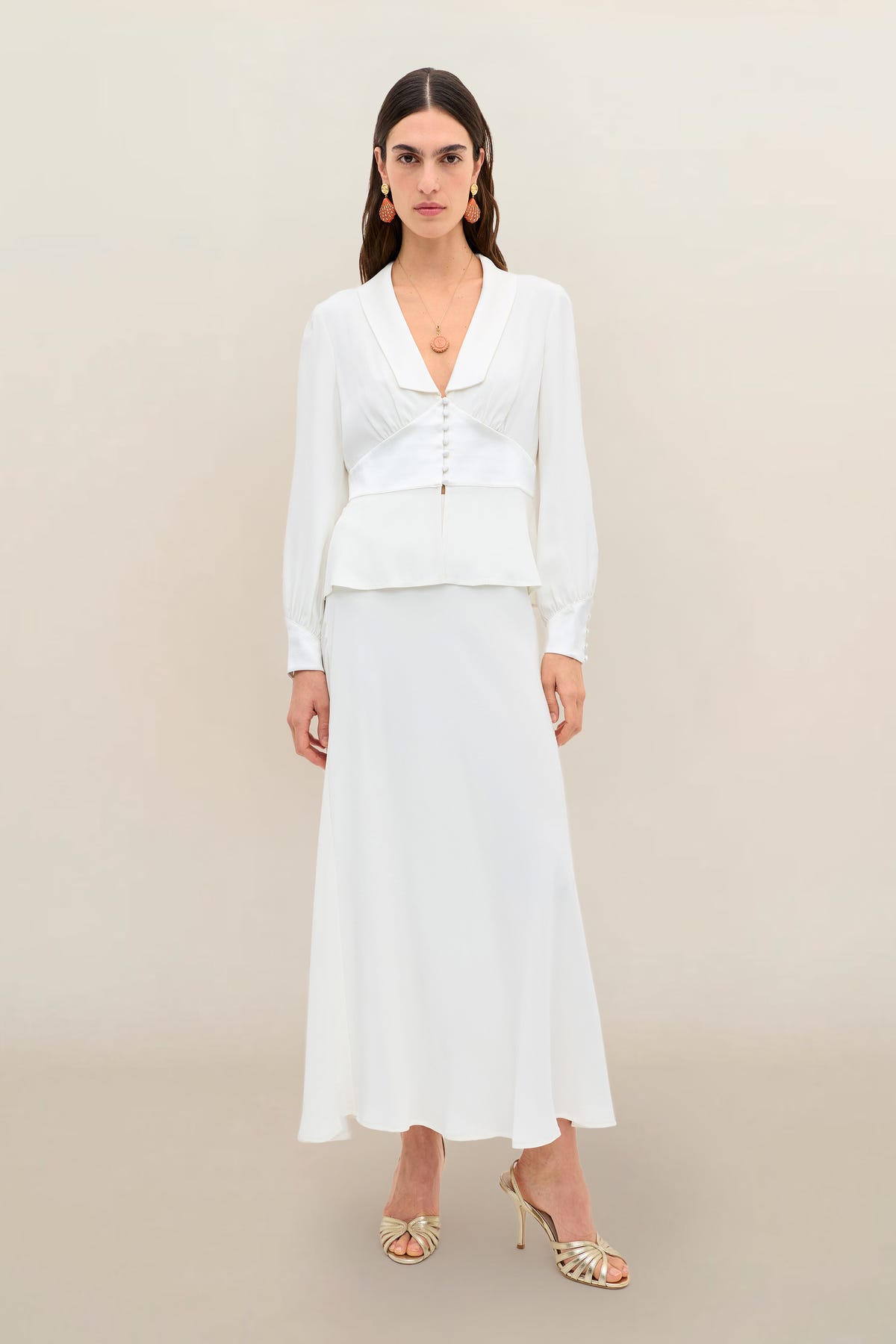 A woman stands against a plain beige background wearing the “Adeline - Ivory,” a long-sleeved silk-crepe blouse with gold buttons and subtle peplum detailing, paired with a matching white maxi skirt. She completes the elegant bridal ensemble with gold strappy heels and gold jewelry, bringing a contemporary twist to this classic look.