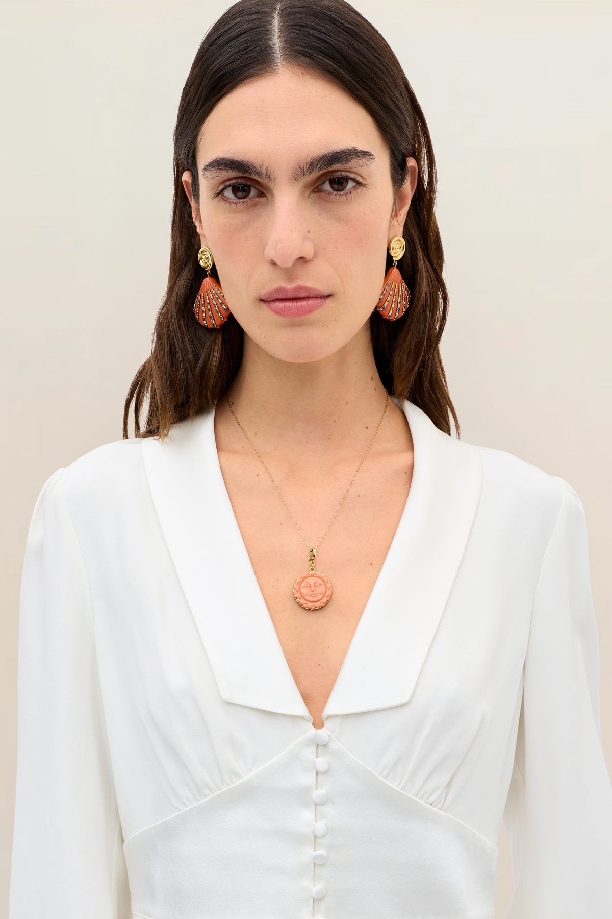 A woman with straight brown hair wears the Adeline - Ivory silk-crepe v-neck blouse, accented by gold and red fan-shaped earrings and a gold necklace with a round pendant. She stands against a plain, light background, looking directly at the camera—the elegance of the Adeline - Ivory bridal top effortlessly complemented by contemporary accessories.