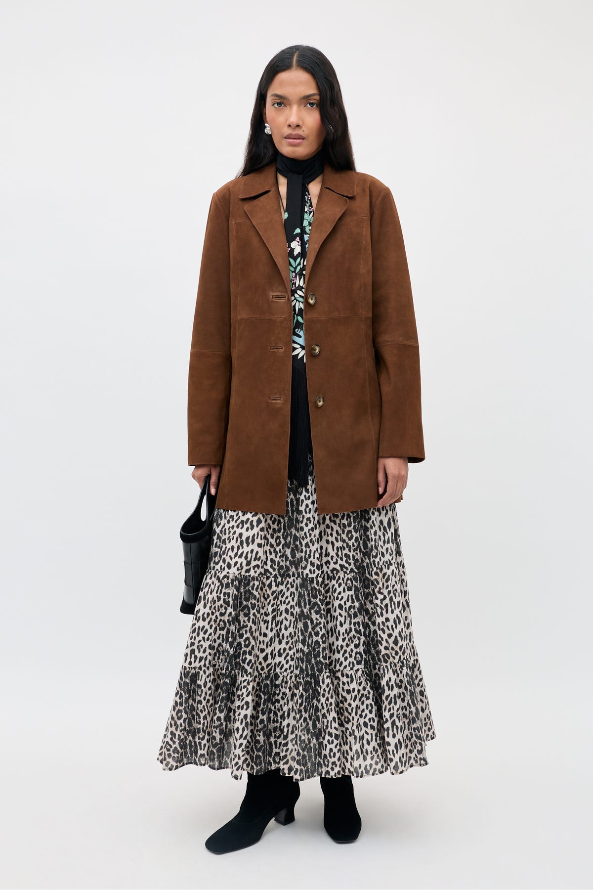 A woman stands facing forward, wearing the Lainey - Chestnut brown suede jacket over a patterned dress with a leopard print skirt, black boots, and holding a small black handbag.