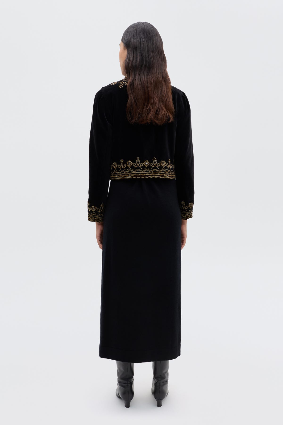 A woman with long brown hair, facing away, wears the Denver - Soutache Black: a black velvet jacket with gold embroidery on the hem and cuffs, over a long black dress and boots. The background is plain light gray.