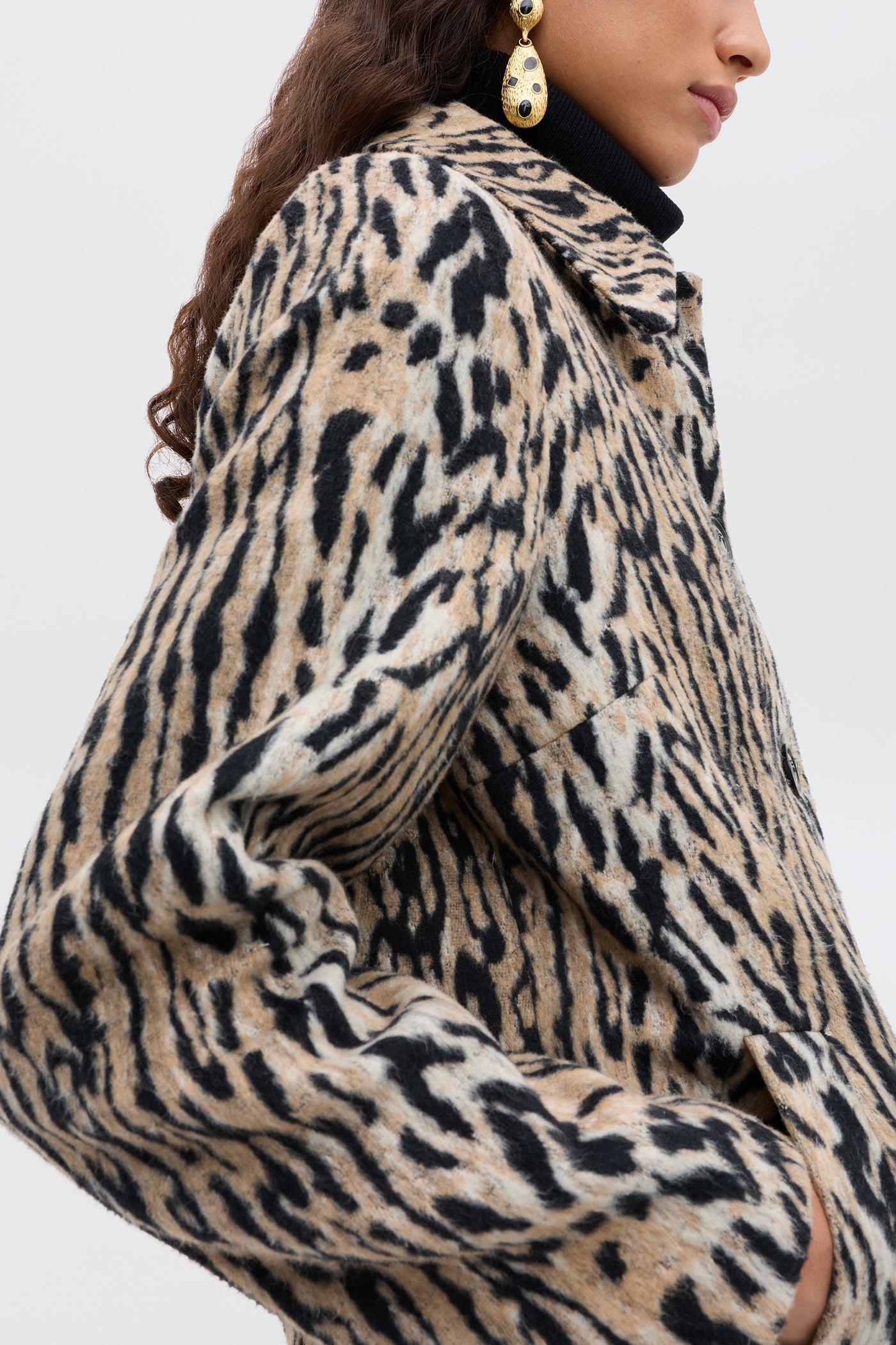 A woman with long curly hair wears the Tommie - Bohemia Leopard jacket, a black turtleneck, and large gold earrings. The image captures her from the shoulders down at a side angle.