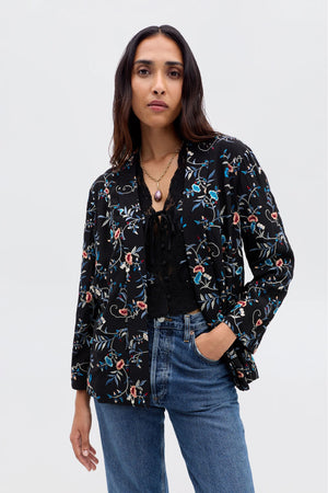A woman wears the Malachy - Botanical Embroidery Black jacket over a black lace top and blue jeans, standing with one hand in her pocket and confidently facing the camera against a plain light background.