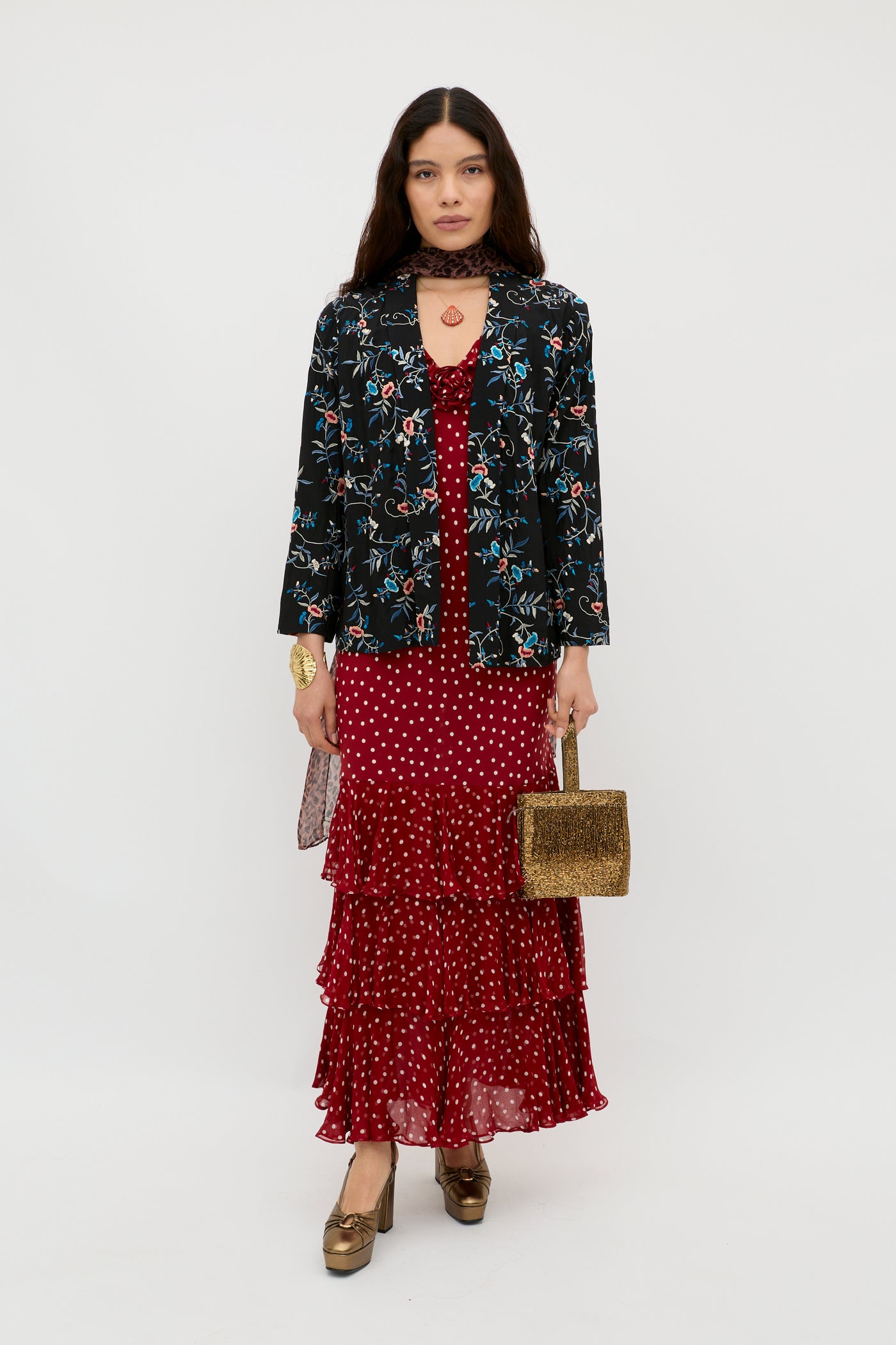 A woman stands against a plain background wearing the Malachy - Botanical Embroidery Black jacket in lightweight crepe over a red polka dot ruffled dress, paired with gold shoes and a gold handbag. Her long hair is down and her expression is neutral.