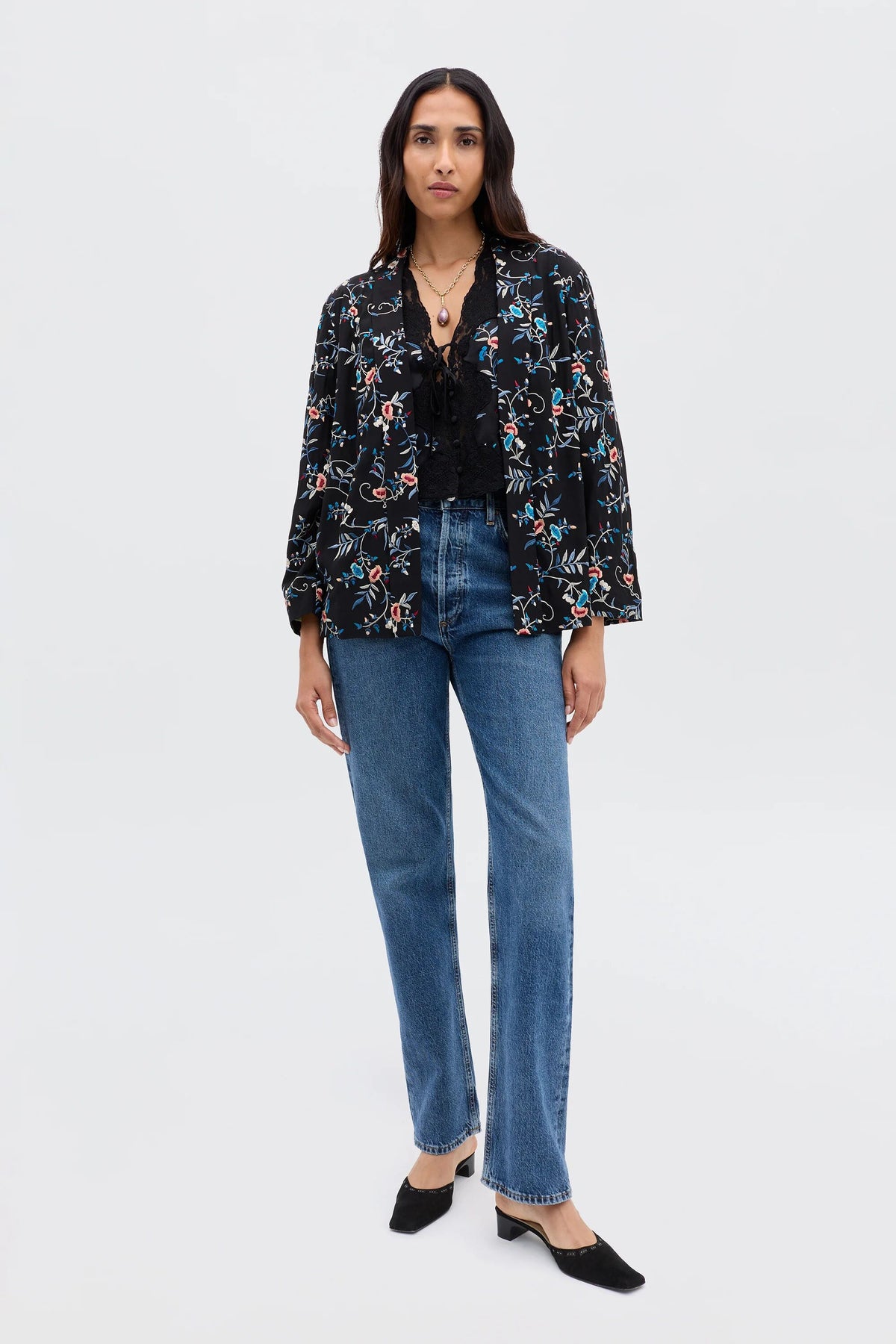 A woman with long dark hair and a neutral expression stands against a plain background wearing the Malachy - Botanical Embroidery Black jacket in lightweight crepe, paired with a black lace top, blue high-waisted jeans, and black heeled mules.