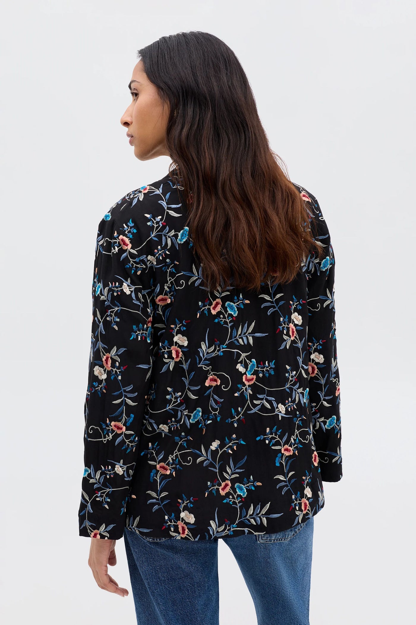 A woman with long, wavy brown hair is seen from behind, wearing the Malachy - Botanical Embroidery Black jacket with blue jeans against a plain light background.