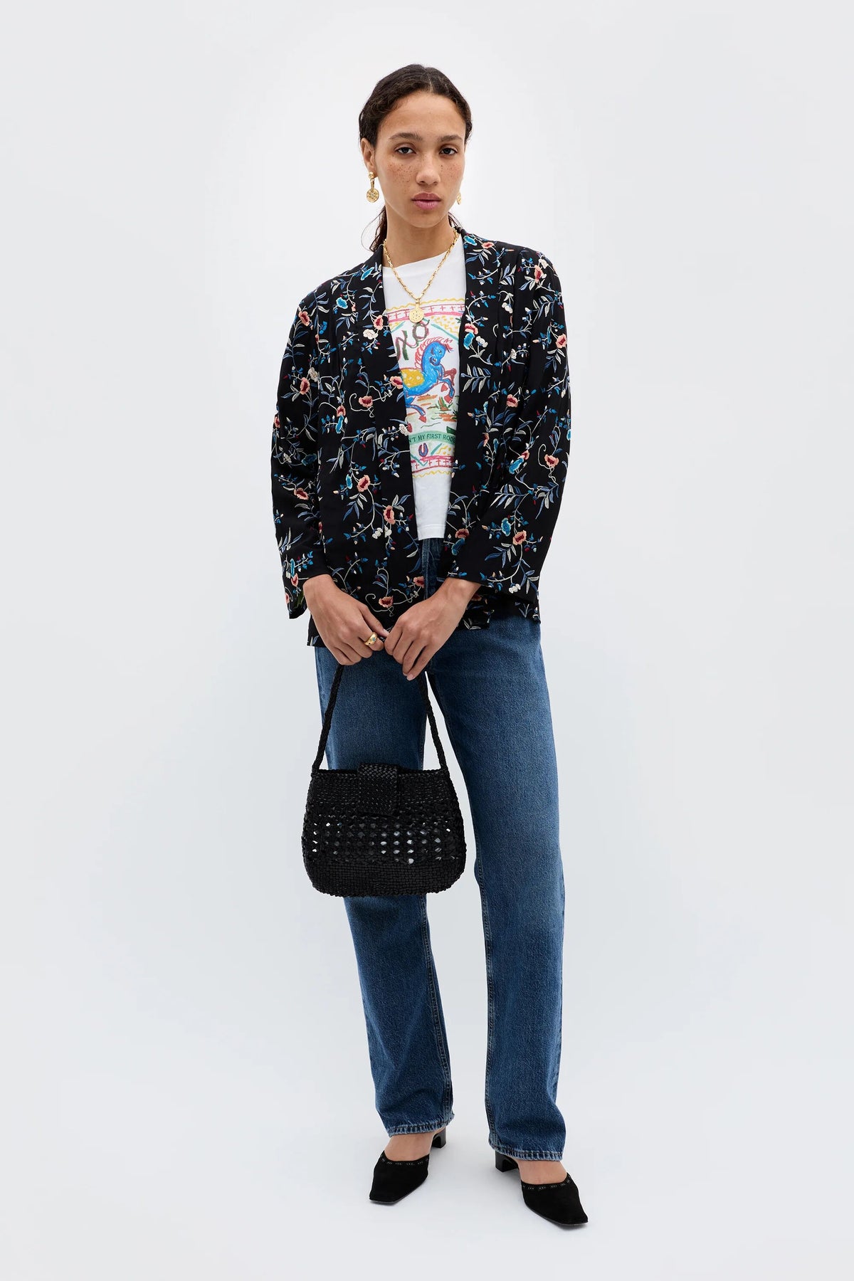 A woman stands against a plain background wearing the Malachy - Botanical Embroidery Black jacket, a colorful graphic T-shirt, blue jeans, black shoes, and holding a black woven handbag.