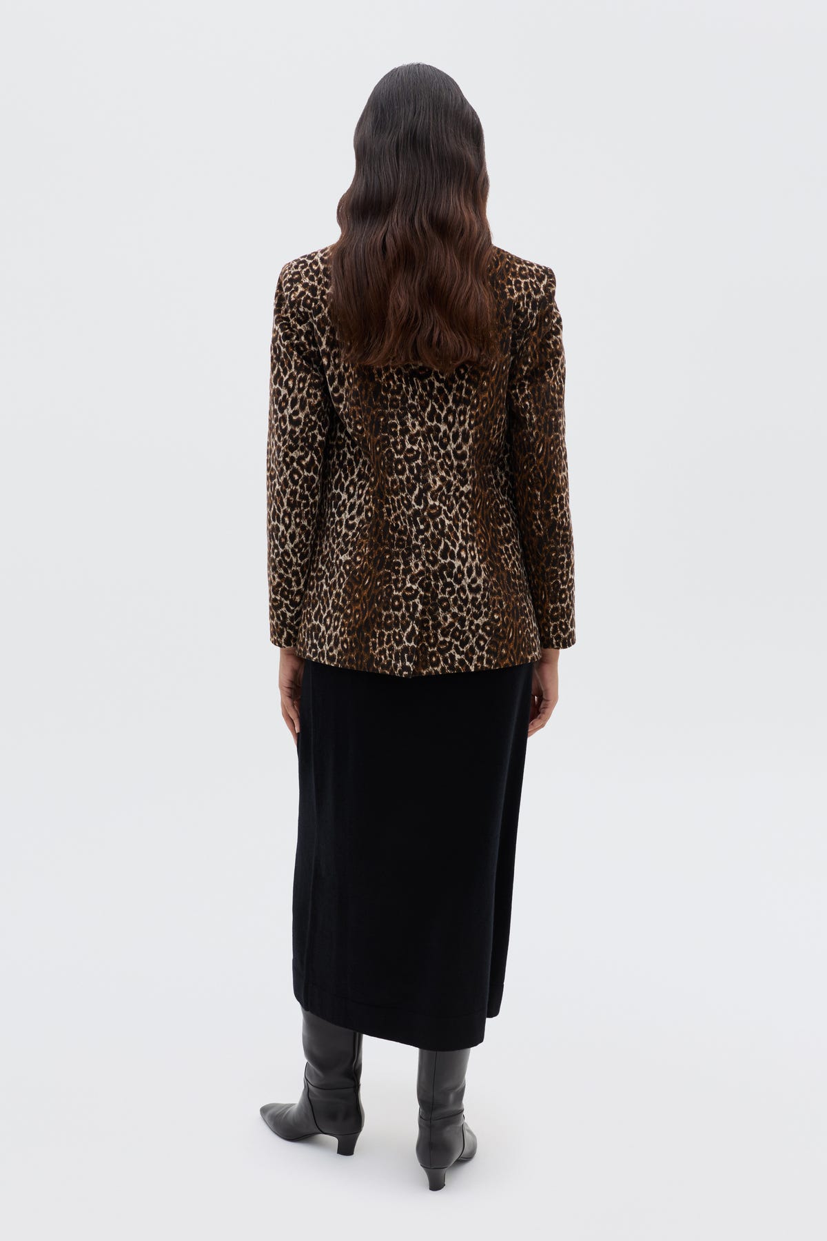A person with long wavy hair stands facing away, wearing the Xander - Lyra Leopard Black jacket, a long black skirt, and knee-high black boots against a plain white background.